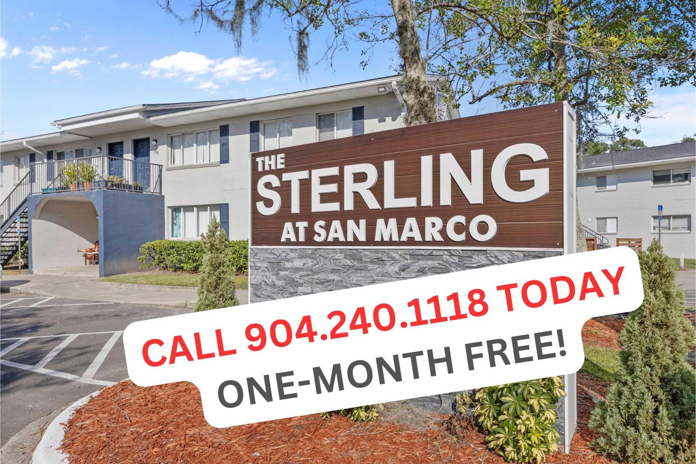 Exterior view of The Sterling at San Marco, showcasing the entrance sign with promotional text, including a phone number and a message about a one-month free offer. Surrounding landscaping features bushes and a clear blue sky in the background.