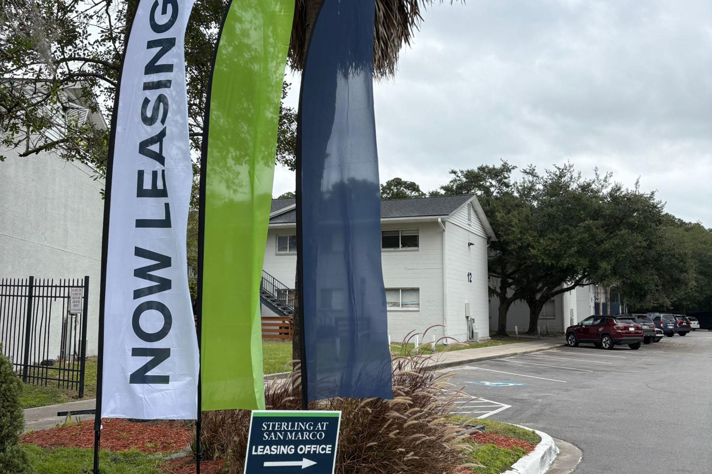 Three tall flags with the text "NOW LEASING" displayed prominently in green and blue, alongside a sign for "STERLING AT SAN MARCO LEASING OFFICE." The setting includes a parking area and nearby buildings, with trees and landscaping in the background.