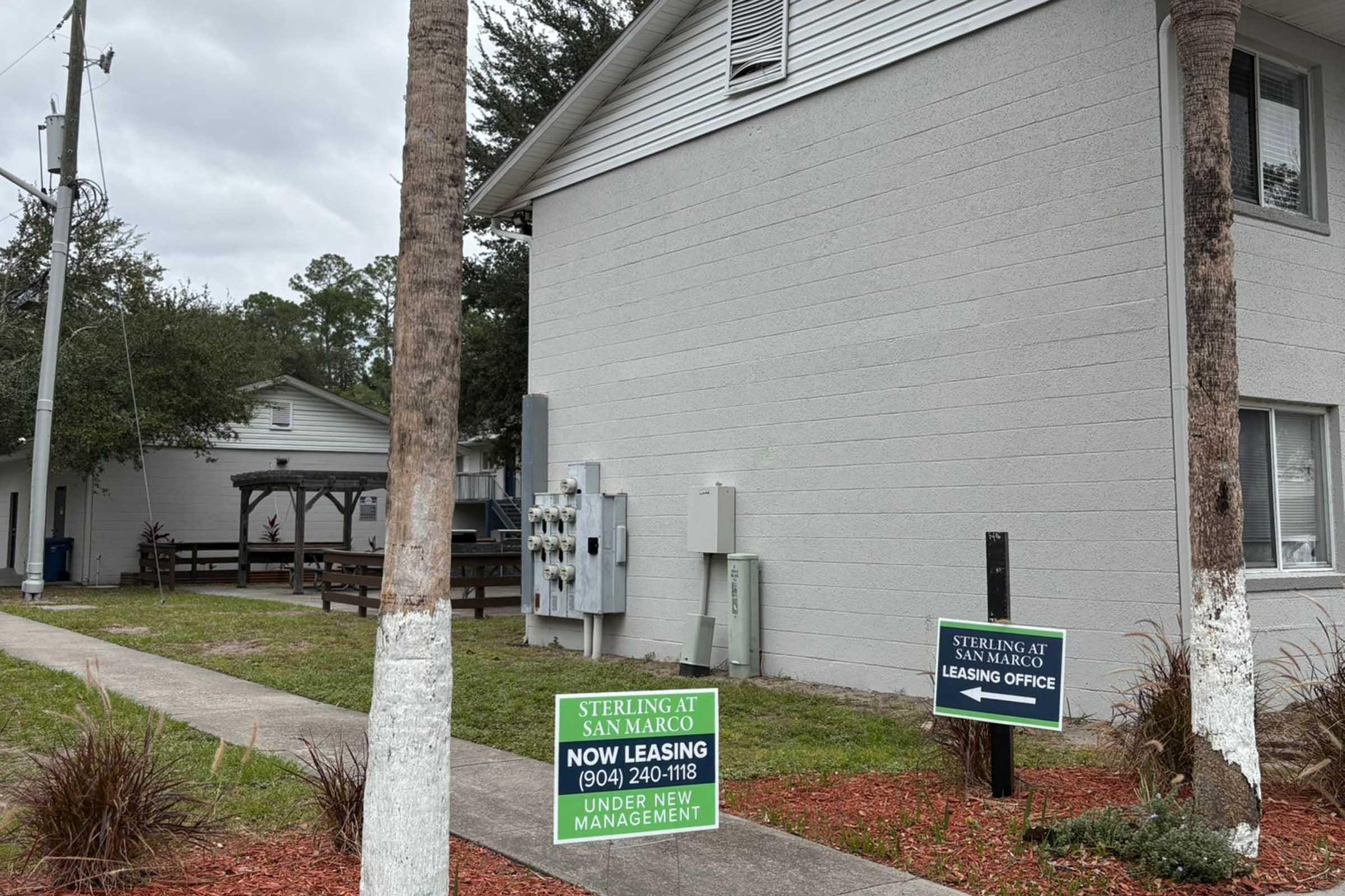 Two signs in front of a residential building. One sign advertises leasing opportunities with the text "NOW LEASING" and a phone number. Another sign indicates "UNDER NEW MANAGEMENT." The building is surrounded by grass and trees, with a sidewalk leading to it.