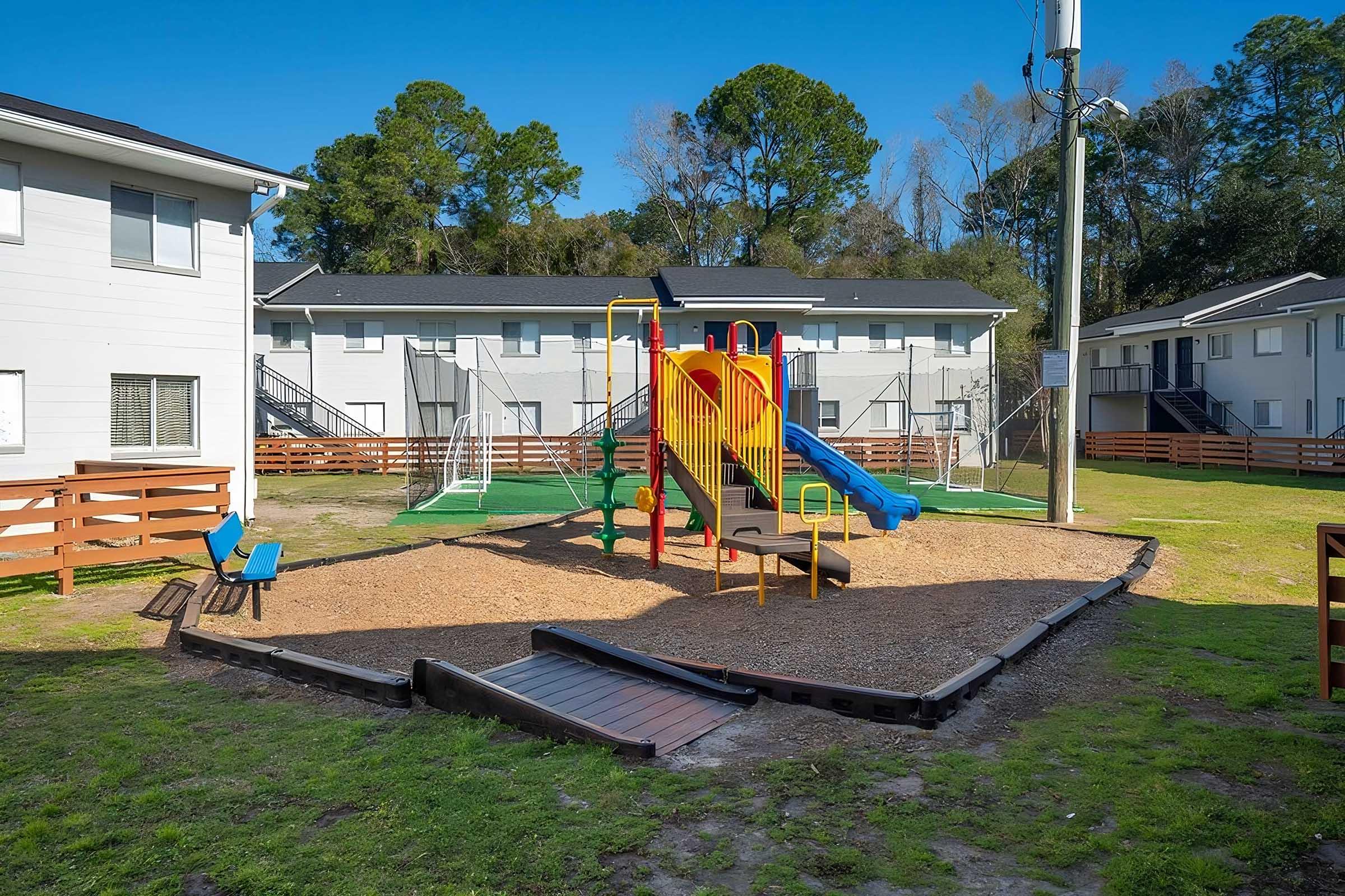 A vibrant playground featuring a slide and climbing structure, surrounded by grassy areas. In the background, there are residential buildings and a soccer field. The scene is sunny, creating a cheerful environment for children to play.