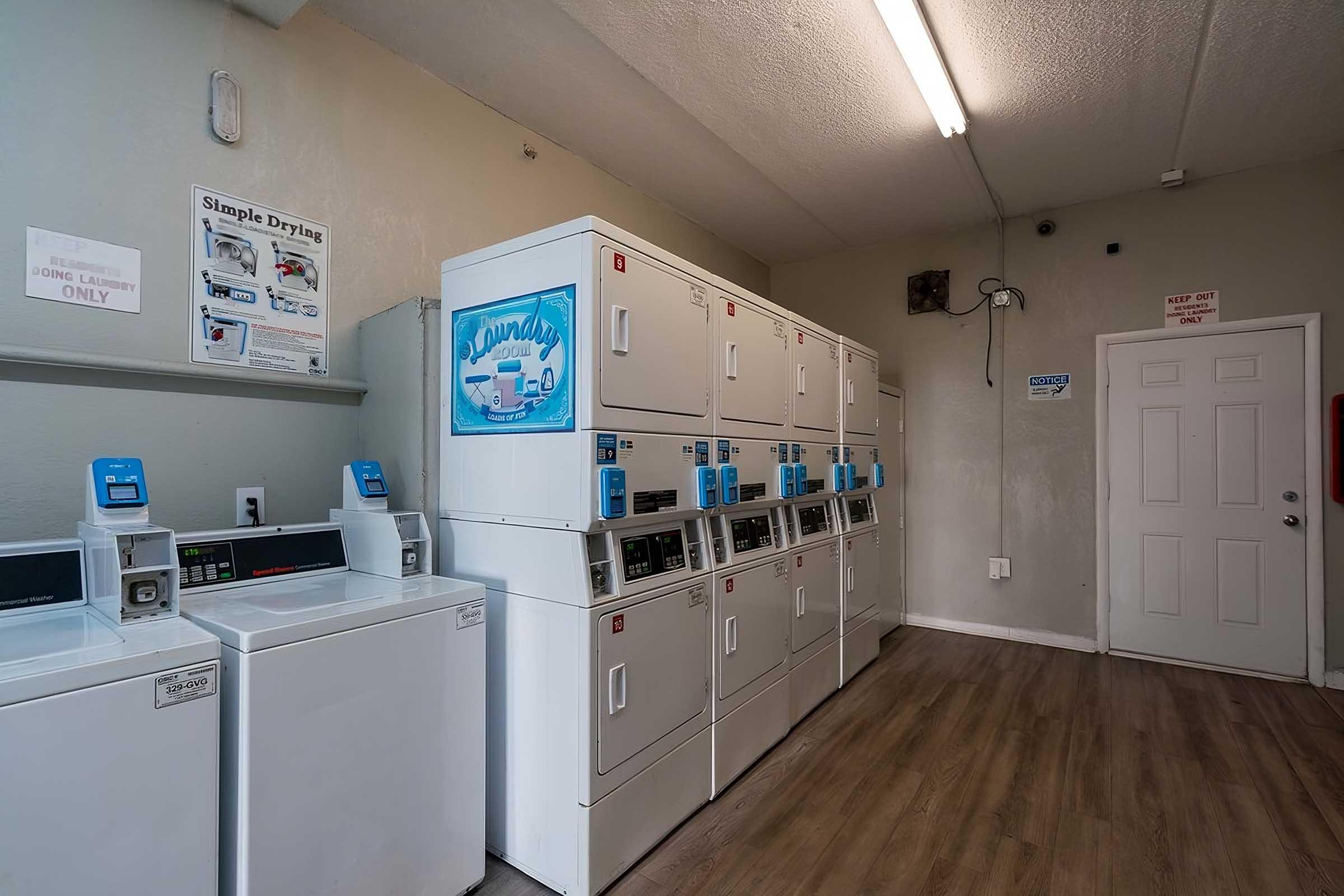 A clean laundry room featuring multiple stacked washing machines and dryers against a wall. There are instructions posted on the wall and a door visible at the end of the room. The flooring is wooden, and the lighting is bright.