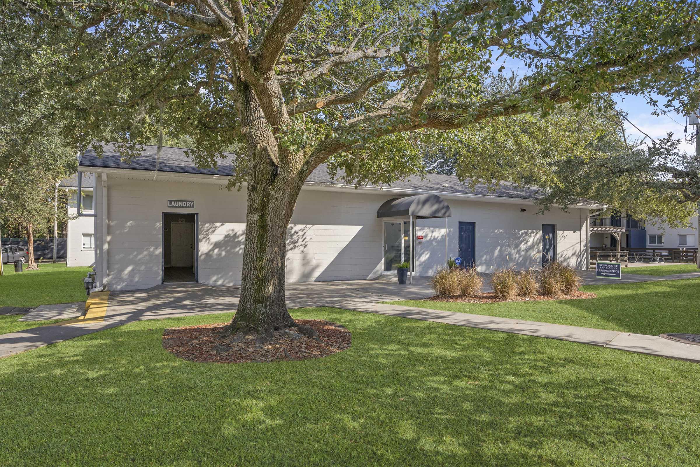 Single-story building with a flat roof, featuring multiple windows and a central entrance. The facade is painted light gray with a dark awning. Surrounding the building is a well-maintained green lawn with a tree providing shade. There are landscaped areas with shrubs in front of the structure.