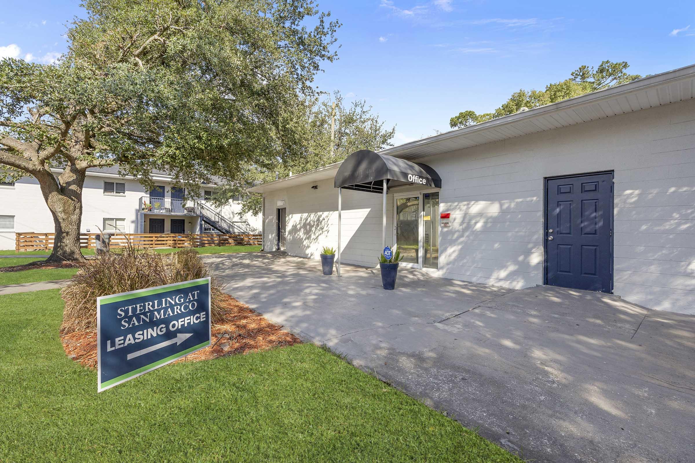 Leasing office exterior of Sterling at San Marco, featuring a covered entrance, flower pots, and a signpost with leasing information. The surrounding area includes green grass and trees, enhancing the welcoming atmosphere of the office space.
