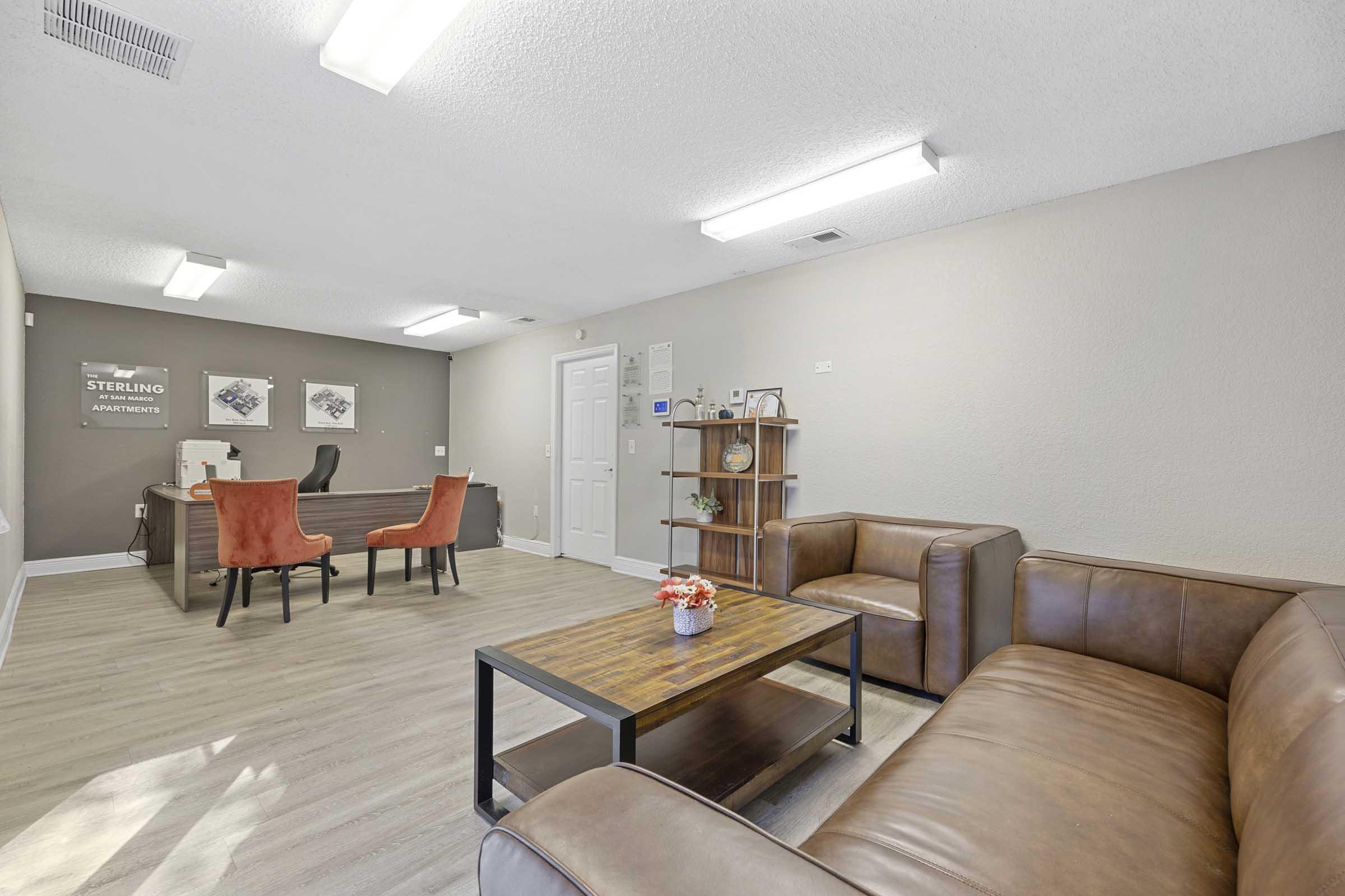 A modern apartment leasing office featuring a reception desk, two orange upholstered chairs, and a brown leather sofa. The space is well-lit with overhead lighting and has a neutral color scheme. A wooden coffee table with a small decorative vase sits in the center, creating a welcoming atmosphere.