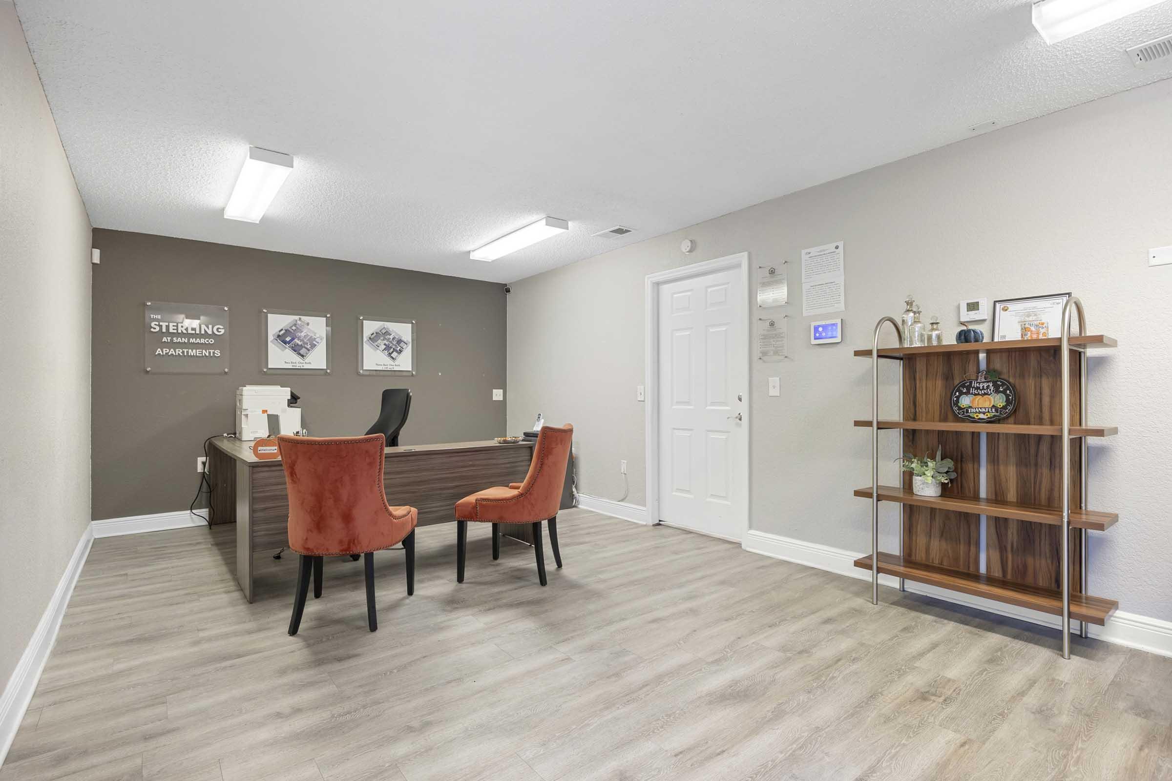 Interior of an apartment office featuring a wooden reception desk, two orange chairs, and a decorative shelf. The walls are painted in neutral tones, and there is modern lighting. Artwork and an information board about apartments are visible, creating a welcoming professional atmosphere.
