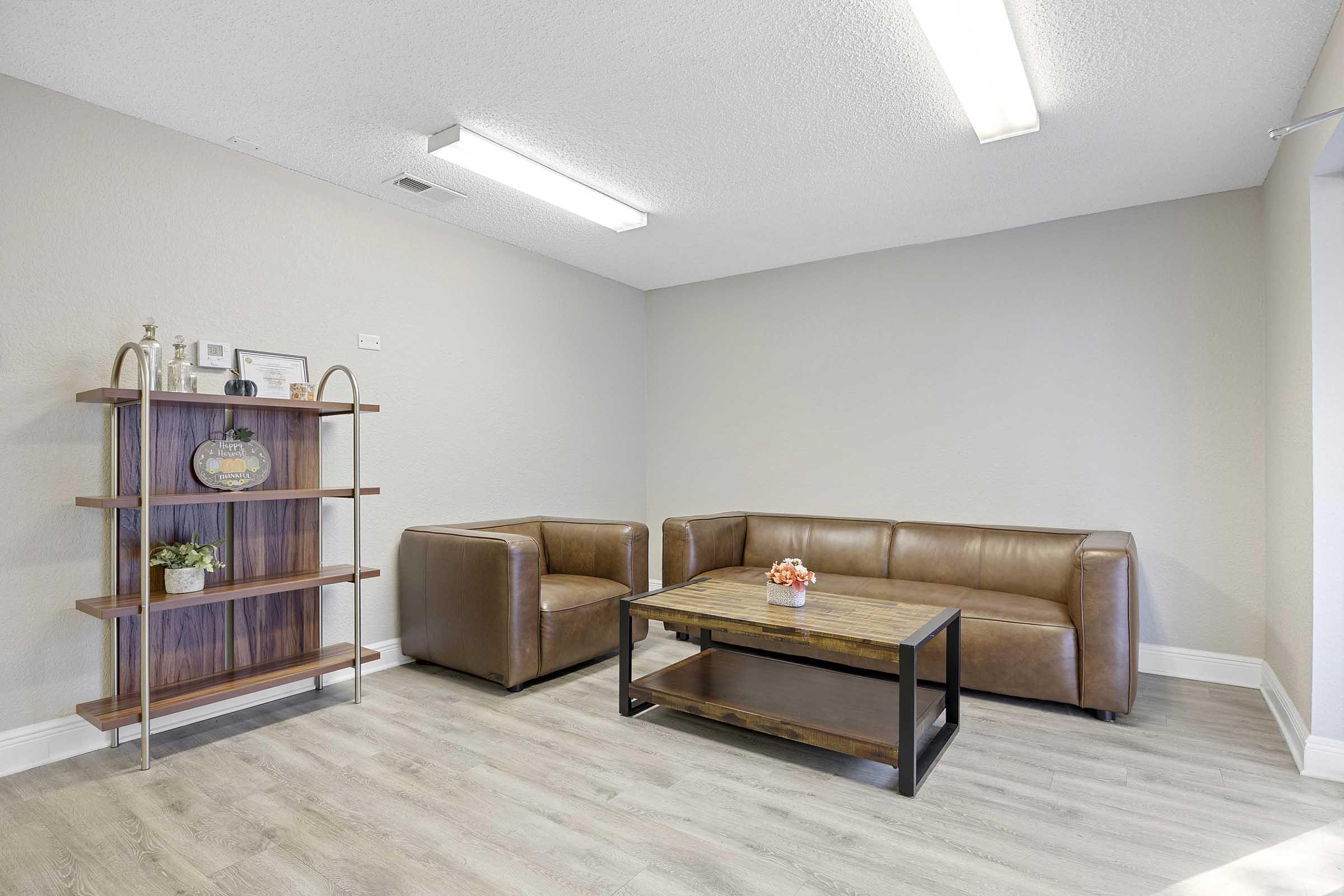 A modern living space featuring a brown leather sofa and an armchair beside a wooden coffee table with a decorative floral arrangement. There’s an open shelf displaying a small plant and framed items against a light gray wall, with bright overhead lighting enhancing the warm atmosphere.