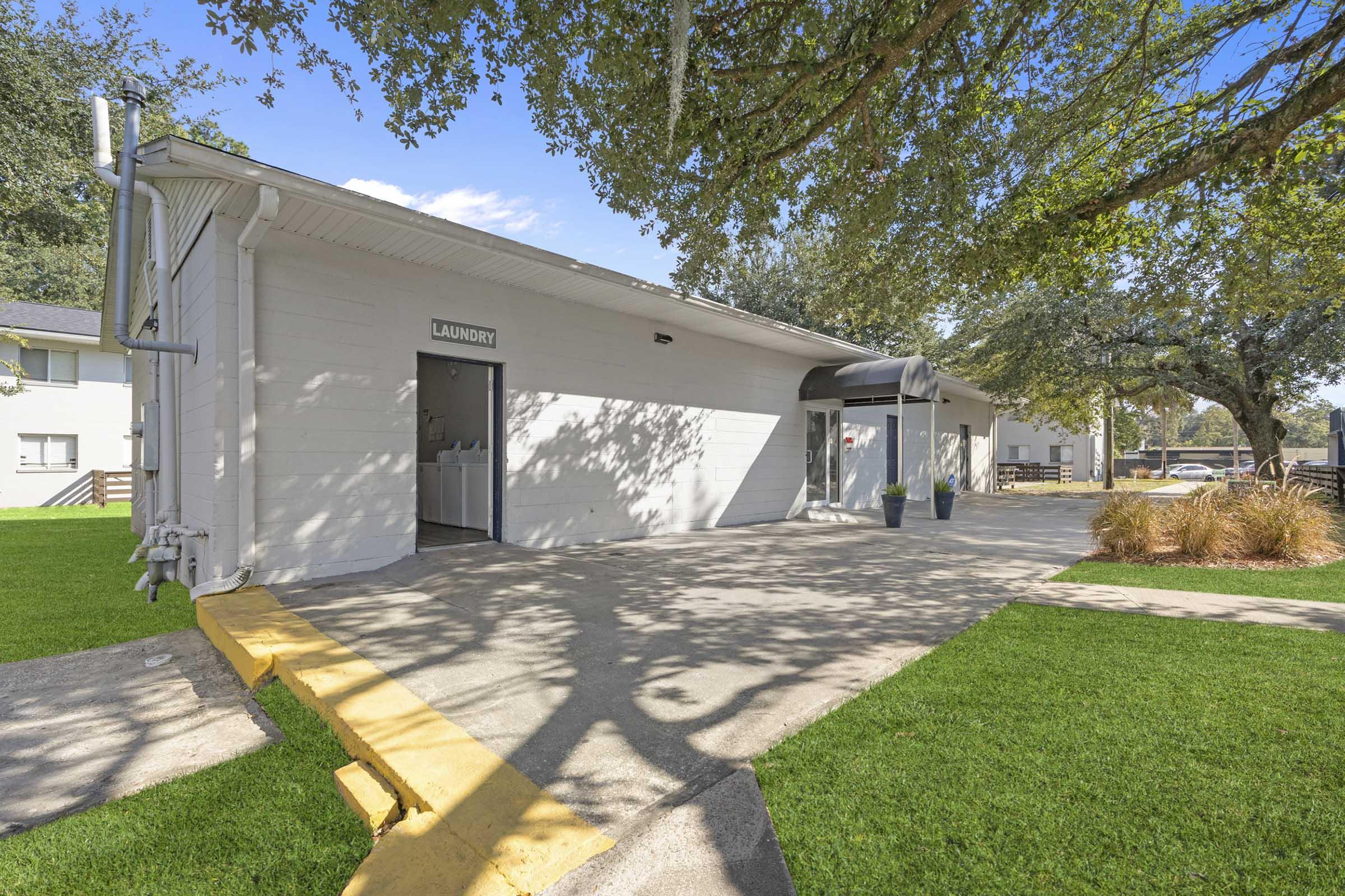 A laundry facility building with a gray exterior, featuring a sign labeled "LAUNDRY." The entrance has large windows and a covered awning. Surrounding the building is well-maintained grass and shrubs, with trees providing shade in the background. The area is sunny and organized.