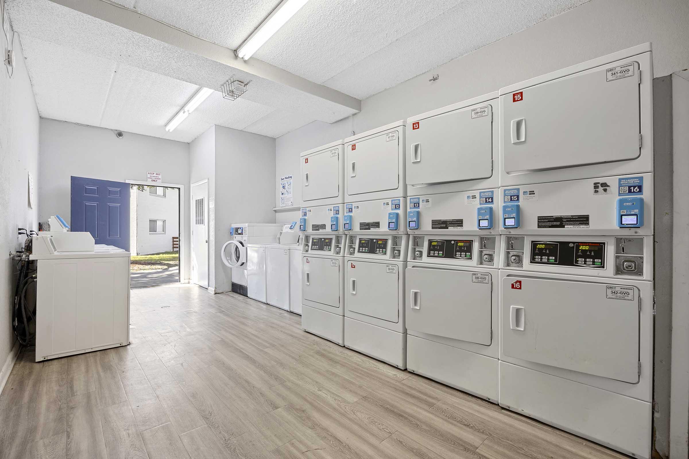 A bright and clean laundry room featuring several stacked washing machines and dryers along one wall. A large door leads to the outside, while another washing machine is positioned at the far end. The space has a modern look with light-colored flooring and organized machines.