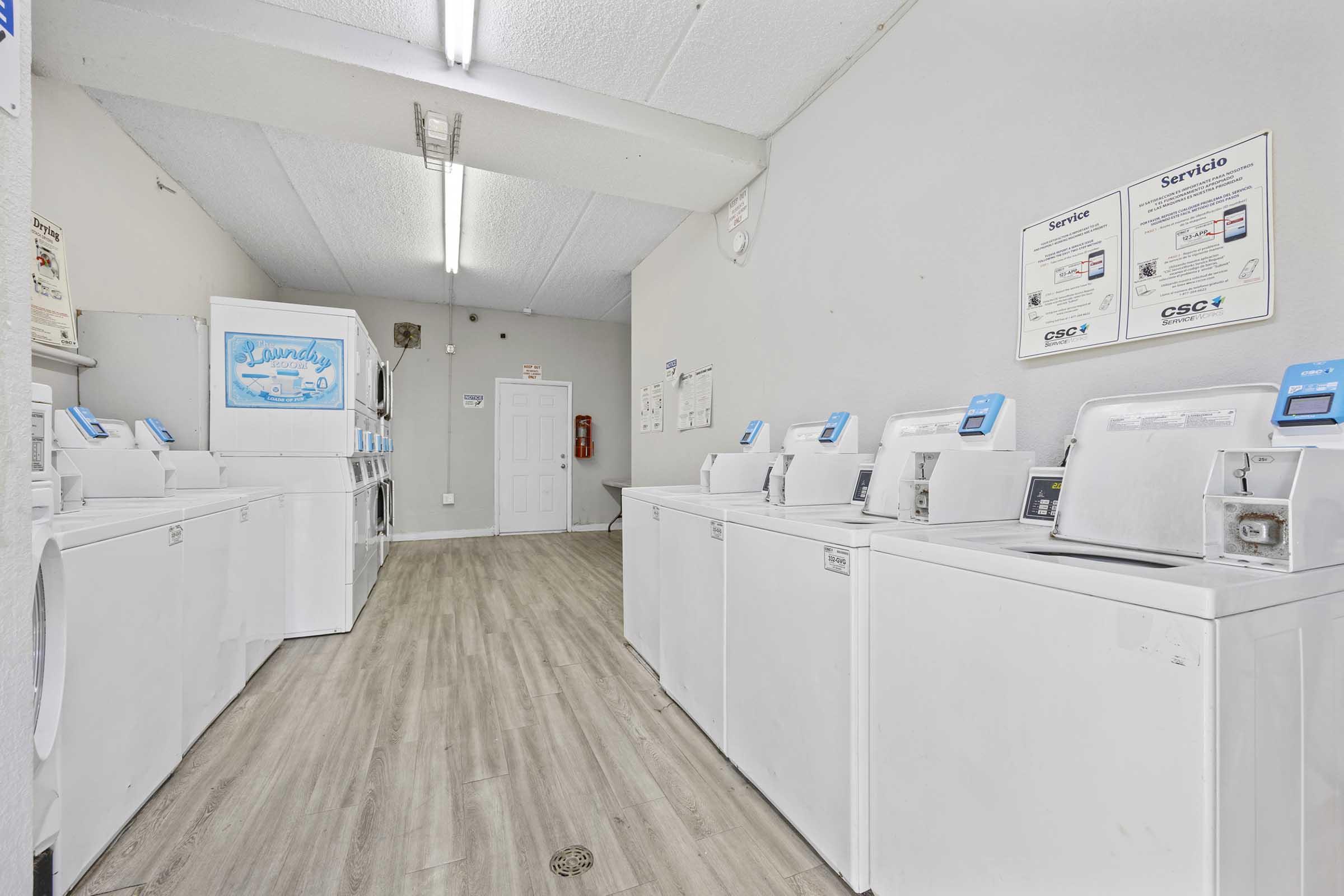 A clean, well-lit laundry room featuring multiple white washing machines and dryers arranged in a row. The walls are painted light gray, and there are service signs displayed. A door can be seen in the background, adding to the functional and organized atmosphere of the space.
