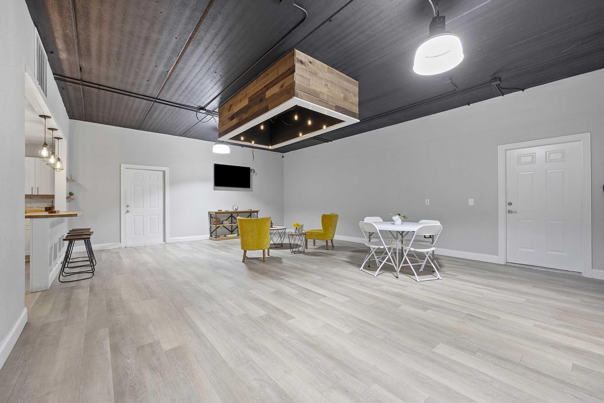 A spacious, modern interior featuring light wooden flooring, a cozy seating area with yellow chairs and a small table, and a dining set with white chairs. The ceiling has a wooden design element, and there's a wall-mounted TV. Soft lighting enhances the inviting atmosphere.