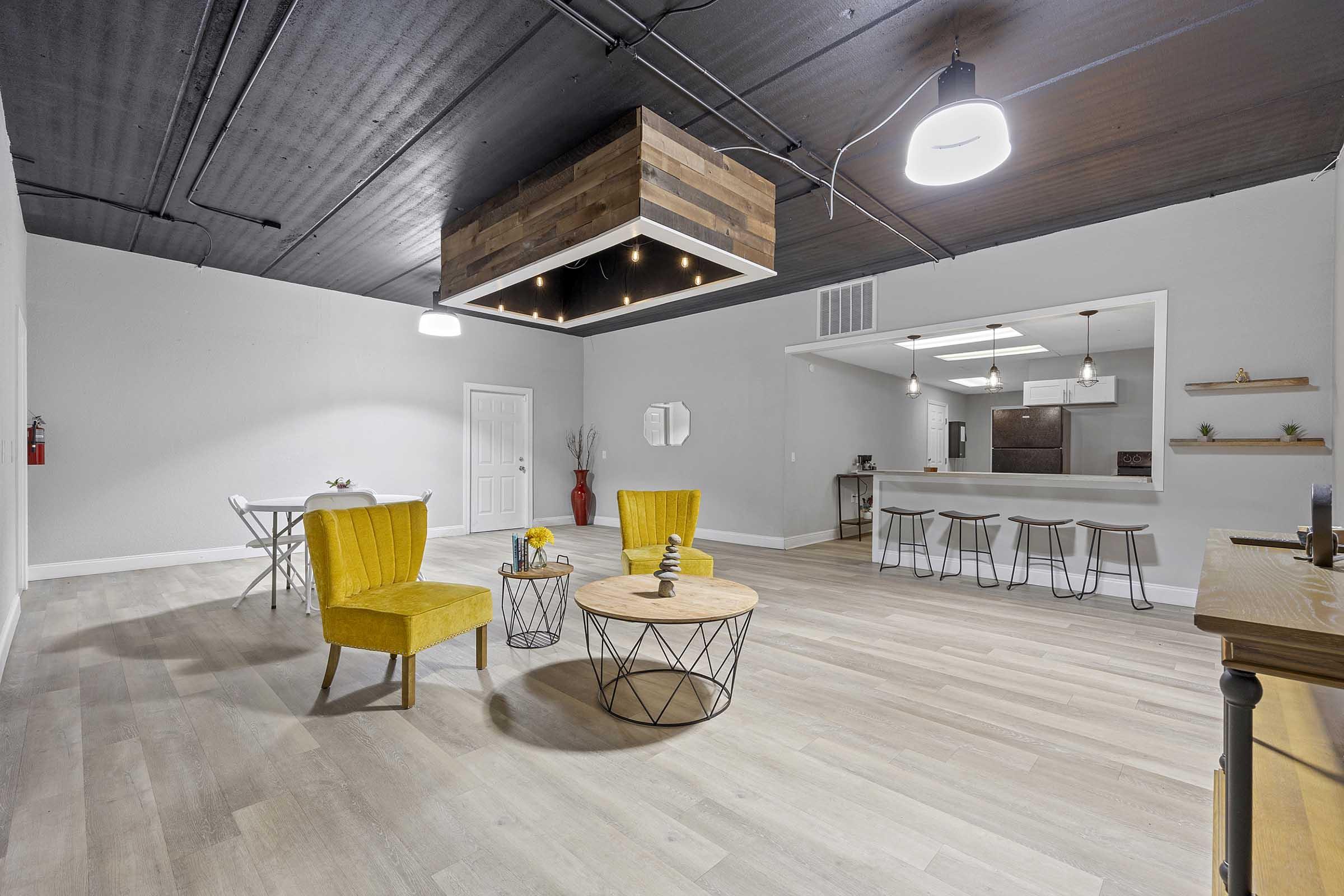 A modern, spacious interior featuring two yellow chairs and a round coffee table in the center. A stylish overhead light fixture hangs above, and the room includes a small dining table in the background. The walls are painted white, with a minimalist design, creating a bright and inviting atmosphere.