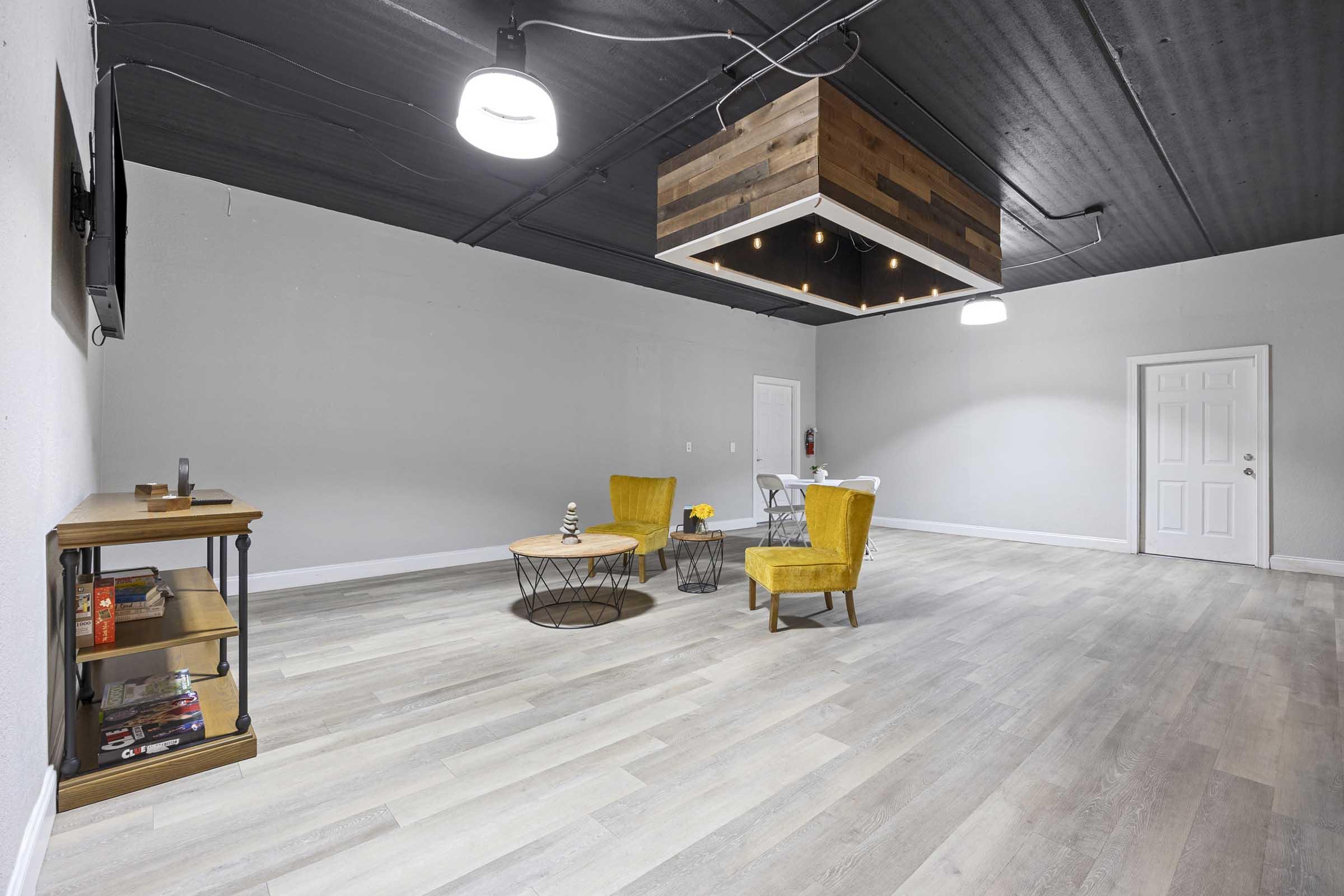A spacious gray room with wooden flooring featuring a cozy seating area. It includes two yellow chairs, a round wooden coffee table, and a small bookshelf with books. A modern light fixture hangs from the ceiling, and a door is visible in the background.
