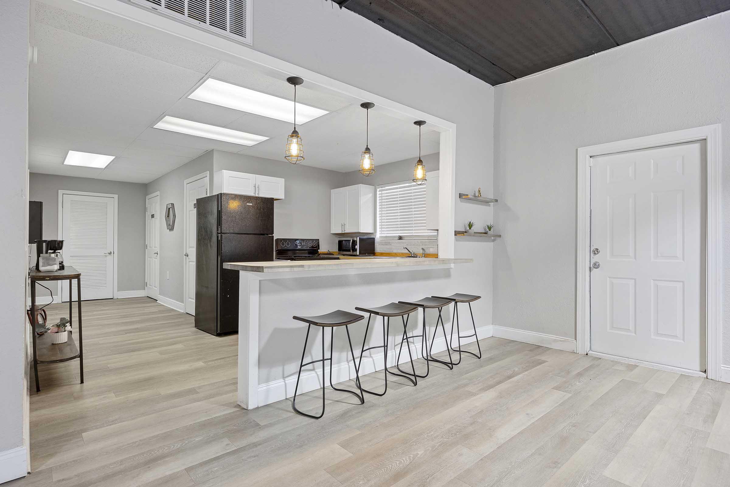 A modern kitchen with a small dining bar featuring four stools. The open layout connects to a bright living area with light-colored walls and wood-style flooring. The kitchen includes black appliances and white cabinets, with pendant lighting above the bar and a door leading to the outside.