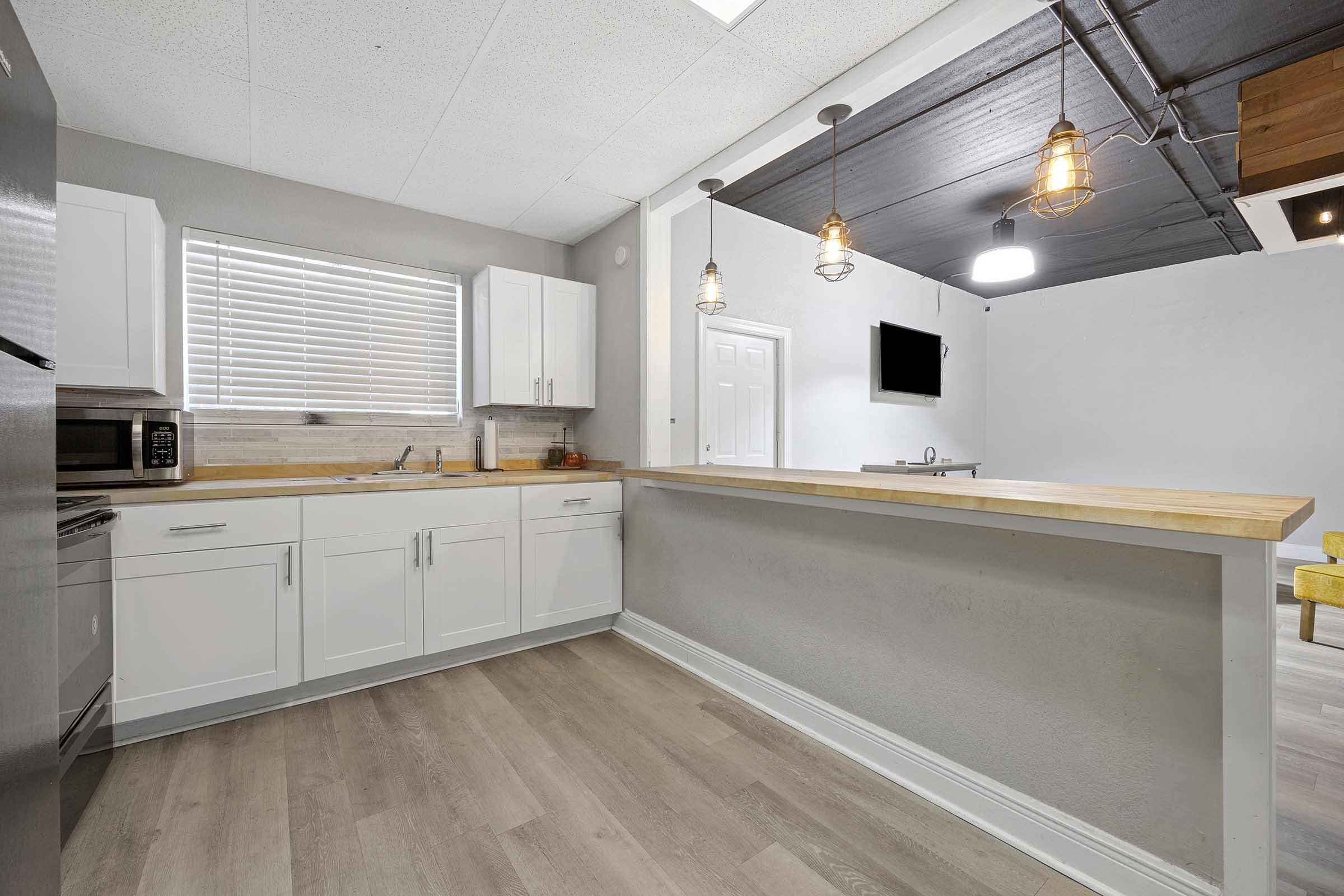 Modern kitchen featuring white cabinets, a wooden countertop, and stainless steel appliances. Natural light filters in through a window with blinds. The open layout includes a bar area with three pendant lights. A wall-mounted TV is visible in the adjoining room, adding a contemporary touch.
