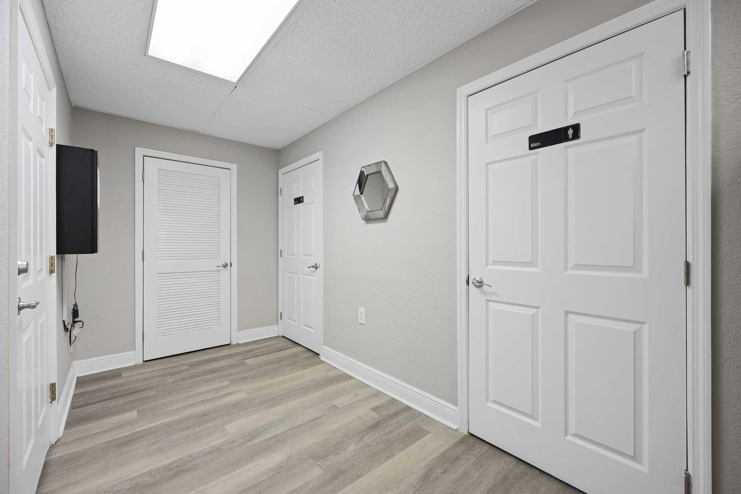 A hallway featuring light-colored walls, wooden flooring, and three white doors. One door is slightly ajar, while a hexagonal mirror hangs on the wall. A small television is mounted on the left wall, and natural light filters in from a skylight above.