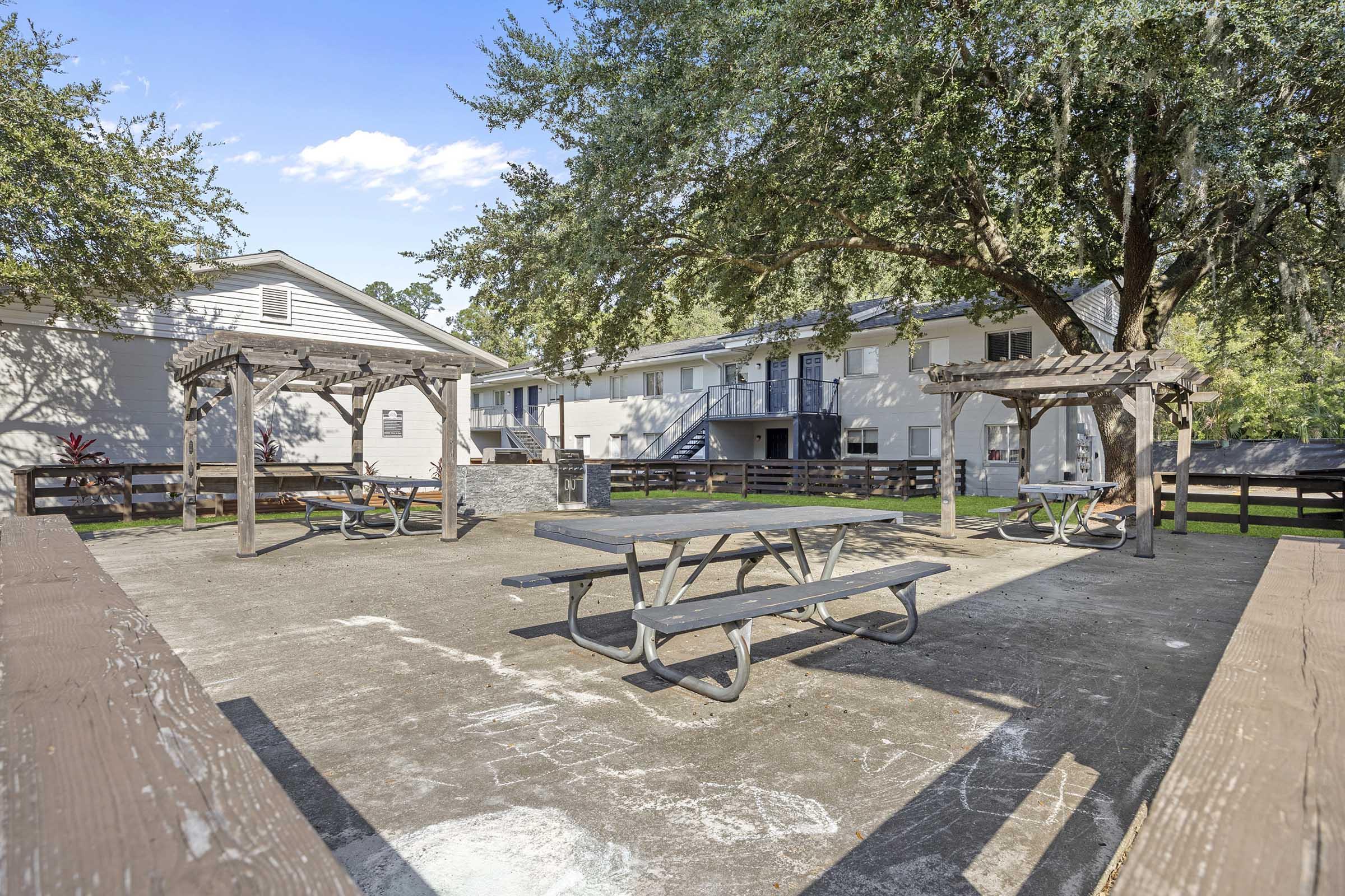 An outdoor communal area featuring several picnic tables beneath shaded wooden pergolas. The concrete space is surrounded by trees and overlooks two multi-story residential buildings in the background under a clear blue sky.