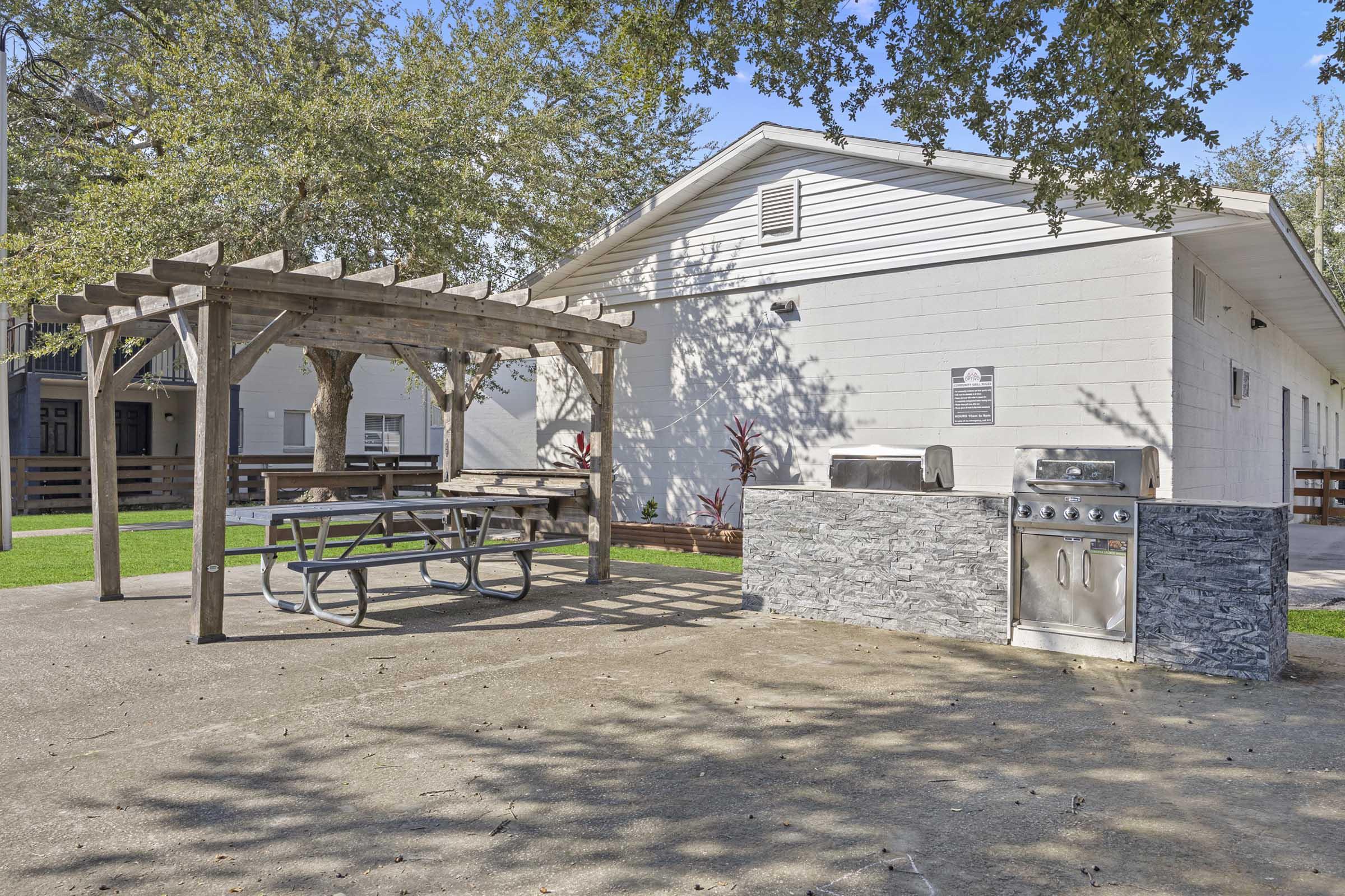 Outdoor barbecue area featuring a stone grill and a covered picnic table under a wooden pergola. The space is surrounded by grass and trees, with a nearby building in the background. The area is well-maintained and designed for social gatherings.