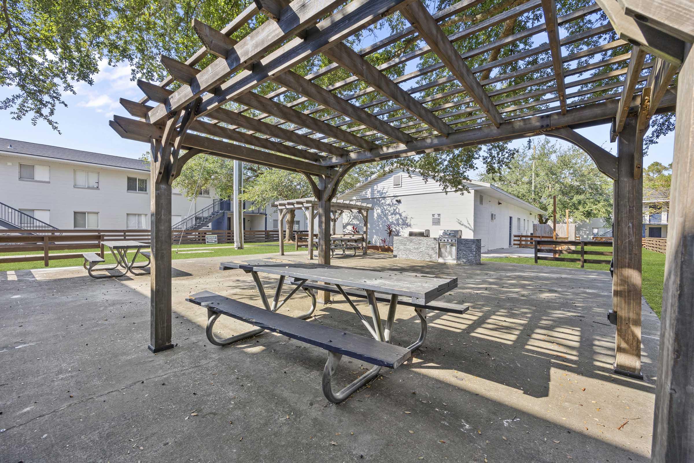 A shaded picnic area featuring a wooden pergola and a picnic table, surrounded by landscaped grass and benches, with nearby residential buildings visible in the background. The setting is bright and inviting, ideal for outdoor gatherings.