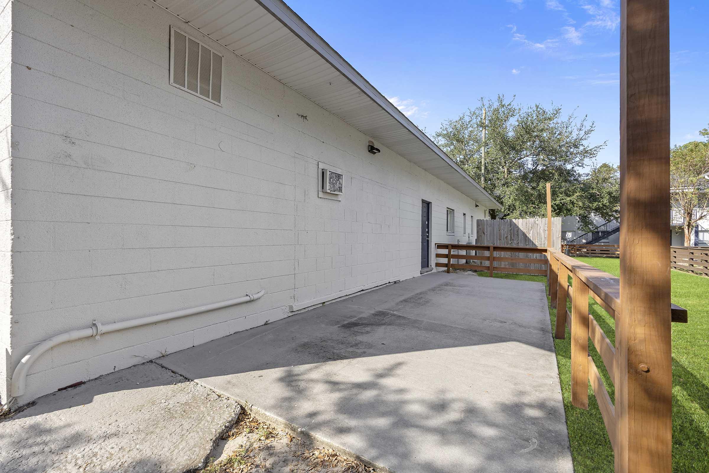 A view of a white exterior wall of a single-story building with a concrete patio in front. There is a wooden railing along one side and a grassy area beyond the patio. The scene is bright with clear blue sky and trees in the background.