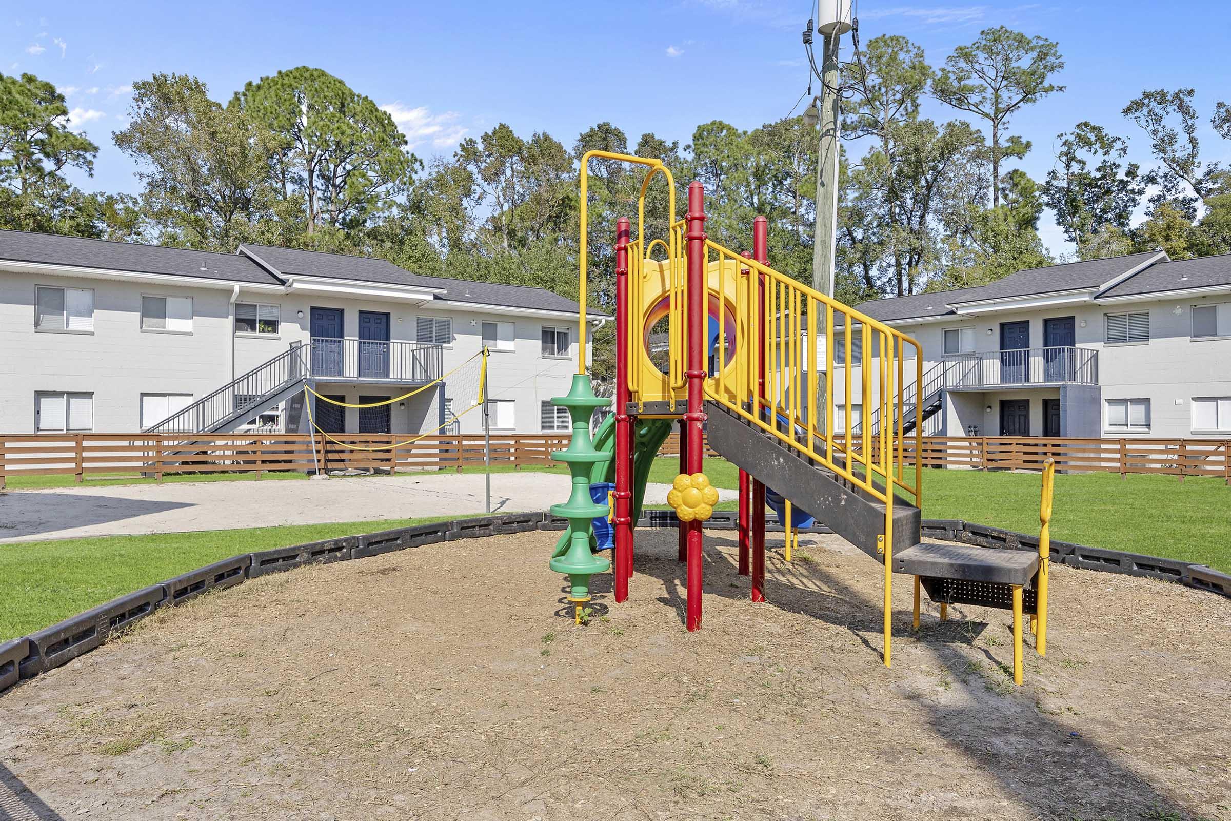 A colorful playground with a slide, climbing structure, and other play equipment, situated in a grassy area surrounded by residential buildings. The scene is sunny, with trees in the background and a clean, inviting environment for children to play.