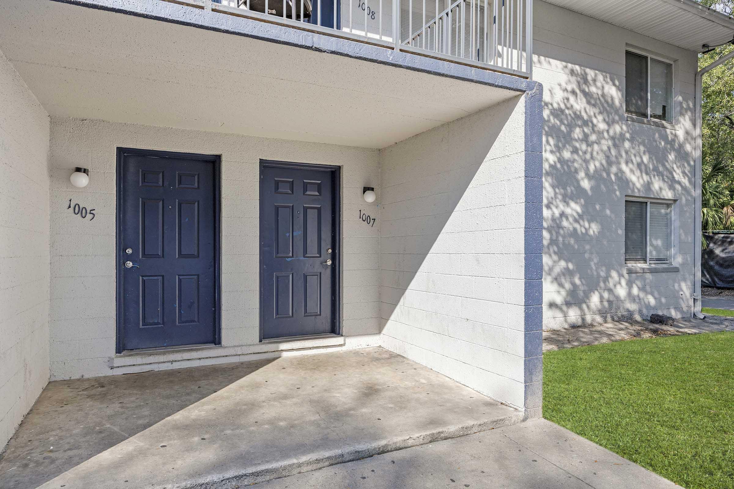 Two identical blue doors labeled "1005" and "1007" are situated at the entrance of a ground-floor apartment building, with a small walkway leading up to them. The exterior features a light-colored wall and a patch of green lawn nearby. Natural light casts shadows on the concrete floor.