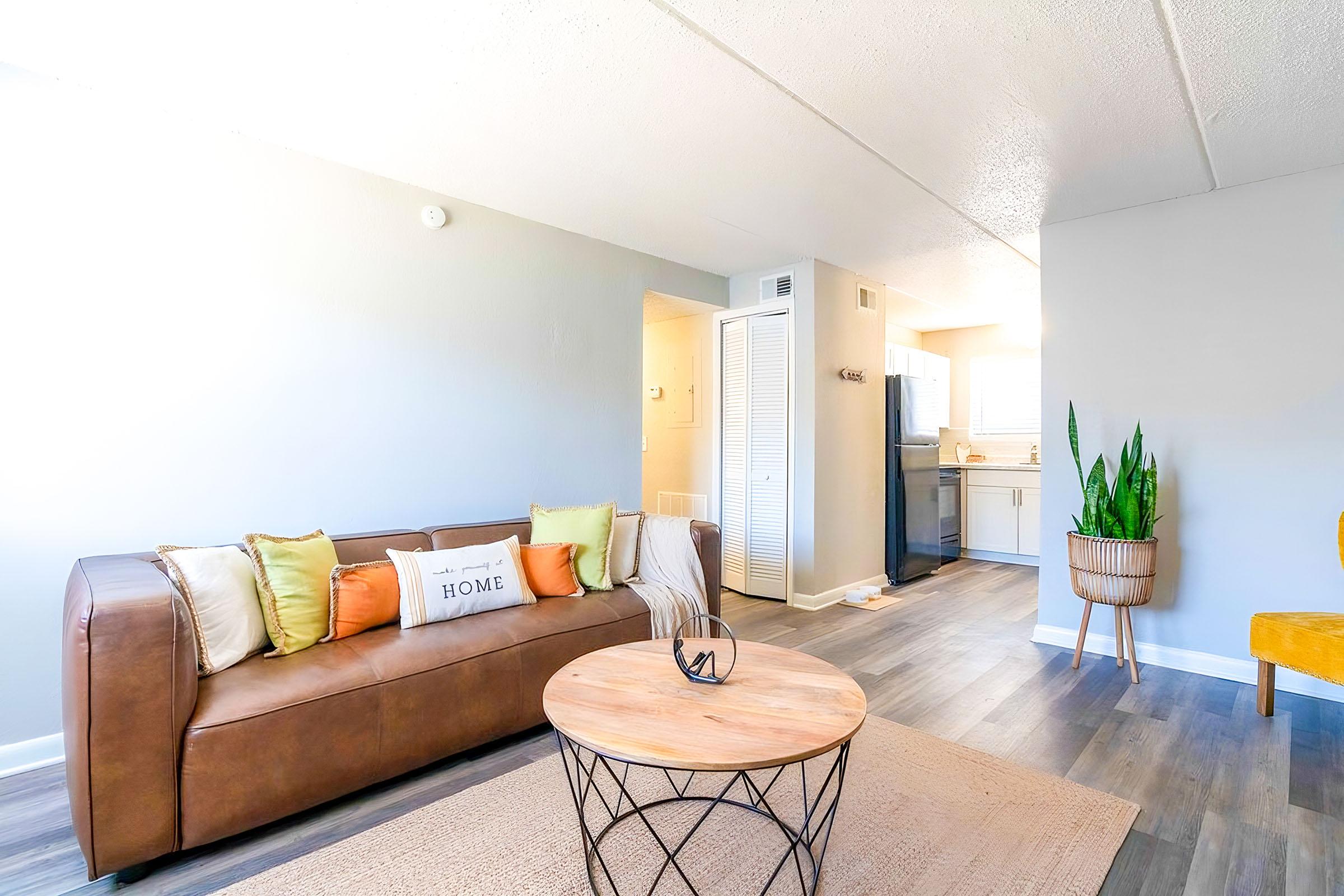 A cozy living room featuring a brown leather sofa adorned with colorful pillows, a round wooden coffee table, and a plant in a woven basket. The space has light-colored walls and a view into a well-lit kitchen area, creating a warm and inviting atmosphere.
