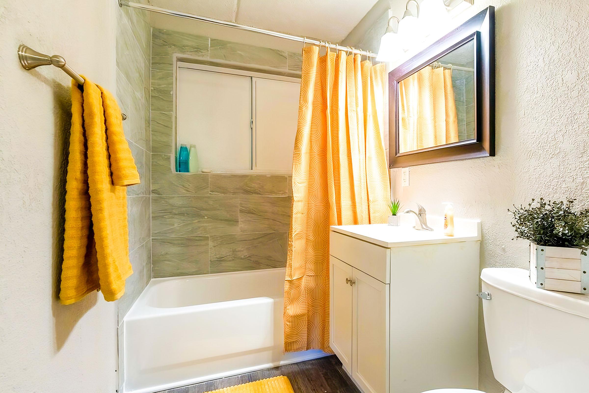 A modern bathroom featuring a bathtub with a glass window, a white vanity with a mirror, and a toilet. Bright yellow curtains and a matching bath mat add color, while a yellow towel hangs on the wall. A small plant and decorative greenery are displayed on the countertop. Natural light illuminates the space.