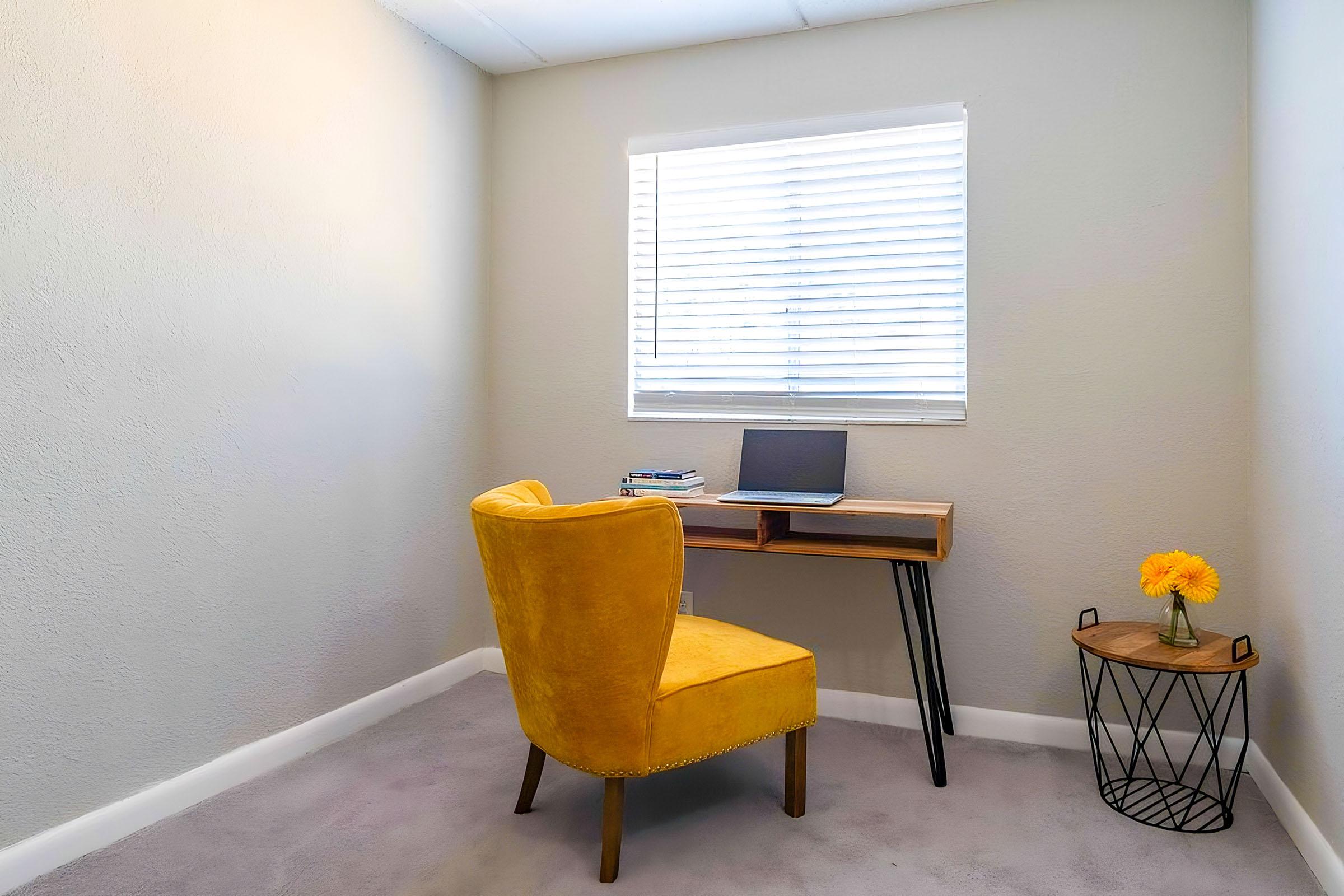 A small home office area featuring a yellow plush chair, a wooden desk with a laptop, and a flower vase on a side table. The space has light gray walls and a window with white blinds, providing natural light. The carpet is light-colored, creating a cozy and inviting atmosphere.