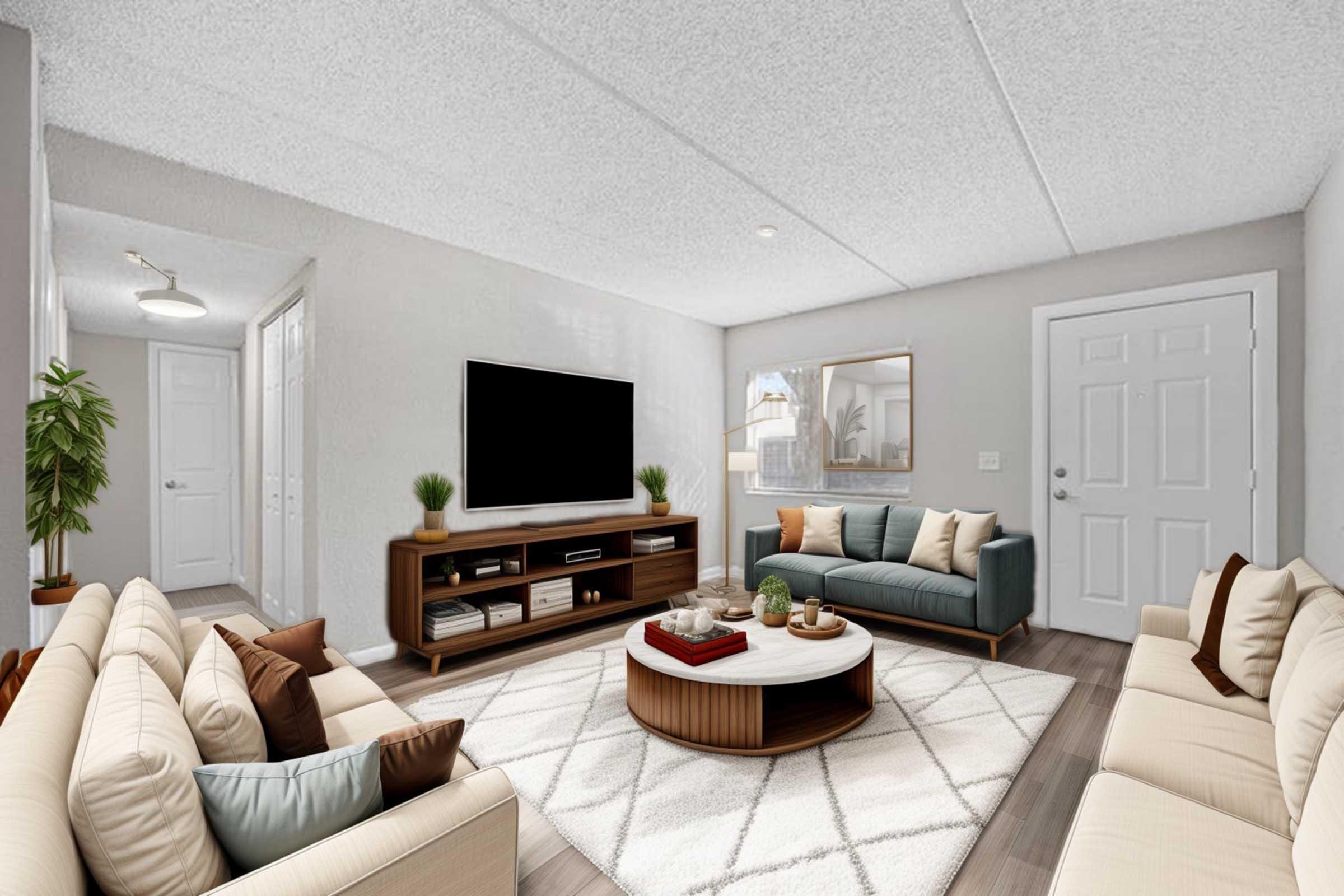 A modern living room featuring light-colored sofas with decorative pillows, a round coffee table with various items, a large TV on a wooden console, and a potted plant. The space has textured ceilings, light walls, and a warm, inviting atmosphere, with natural light coming in from a nearby window.