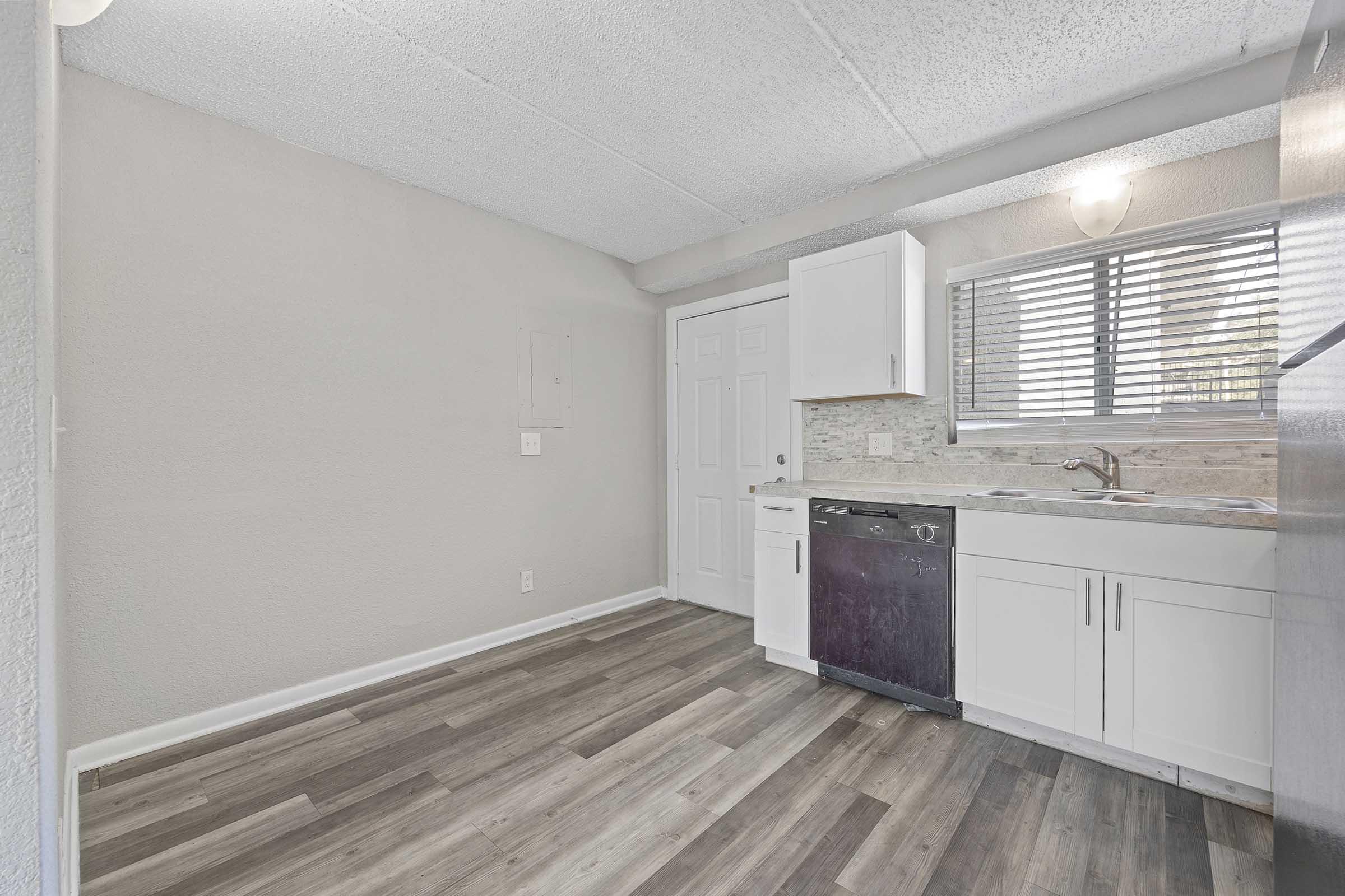 A modern kitchen space featuring light-colored walls, a window with blinds, and white cabinetry. The kitchen includes a sink, dishwasher, and countertops with a stone backsplash. The floor is made of wood-like laminate, creating a warm and inviting atmosphere.