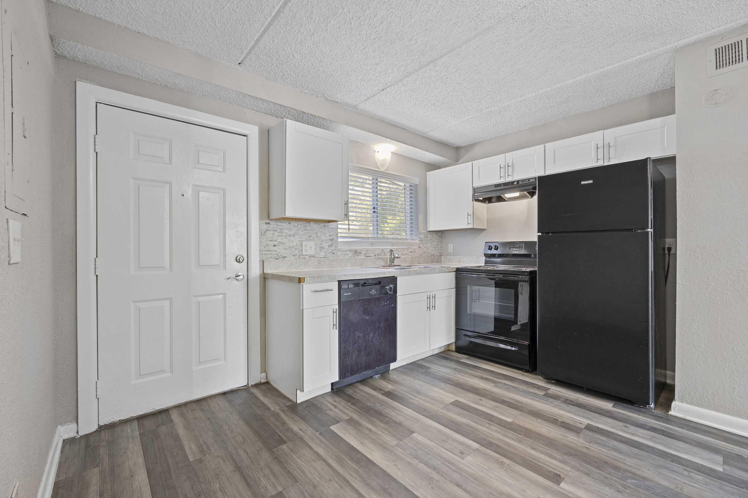 A modern kitchen featuring white cabinetry, a black refrigerator, and an oven. There is a dishwasher beside the sink, with a window allowing natural light. The flooring is a blend of light and dark wood tones, and the walls are painted a neutral color, creating a bright and inviting space.