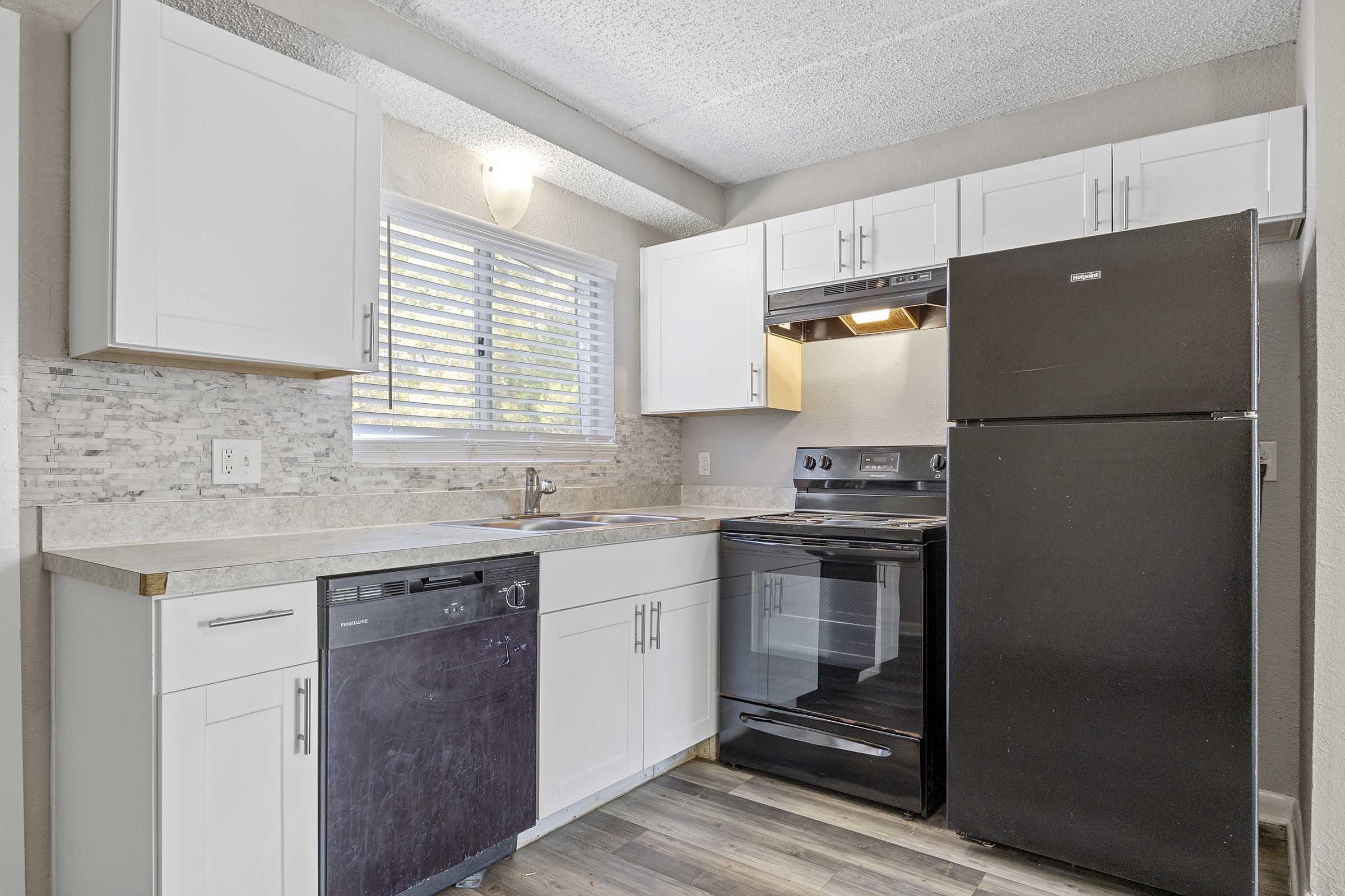 Modern kitchen featuring white cabinetry, a dark refrigerator, and a black oven. The countertop is light-colored with a mix of textures, and there's a window providing natural light. A dishwasher is built into the cabinetry, and the floor is a dark wood-look laminate.