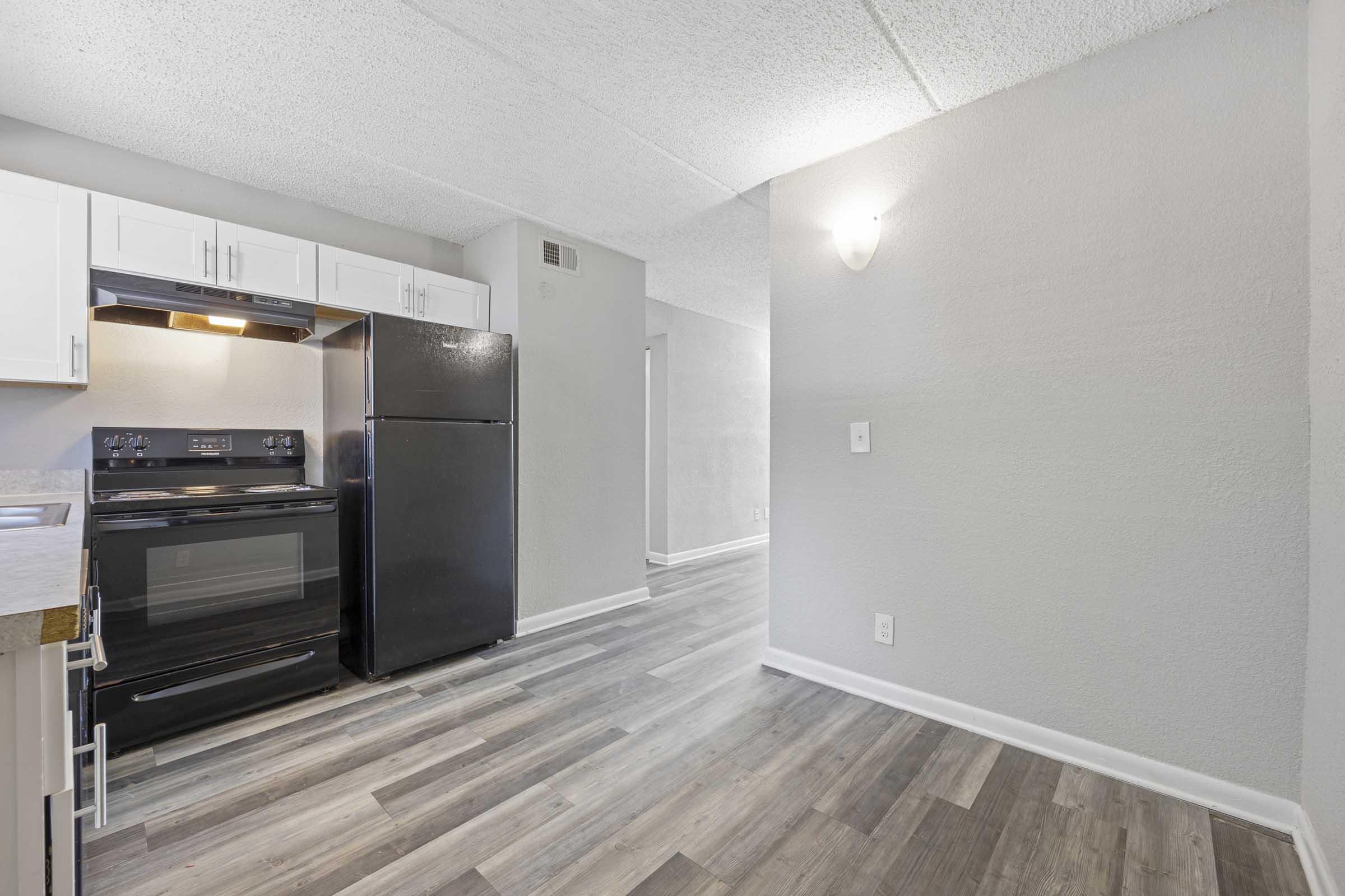 Modern kitchen featuring black appliances, including an oven and refrigerator, with white cabinetry and a gray wall. The space has light-colored laminate flooring and a well-lit area, showcasing an open layout leading into a living space.