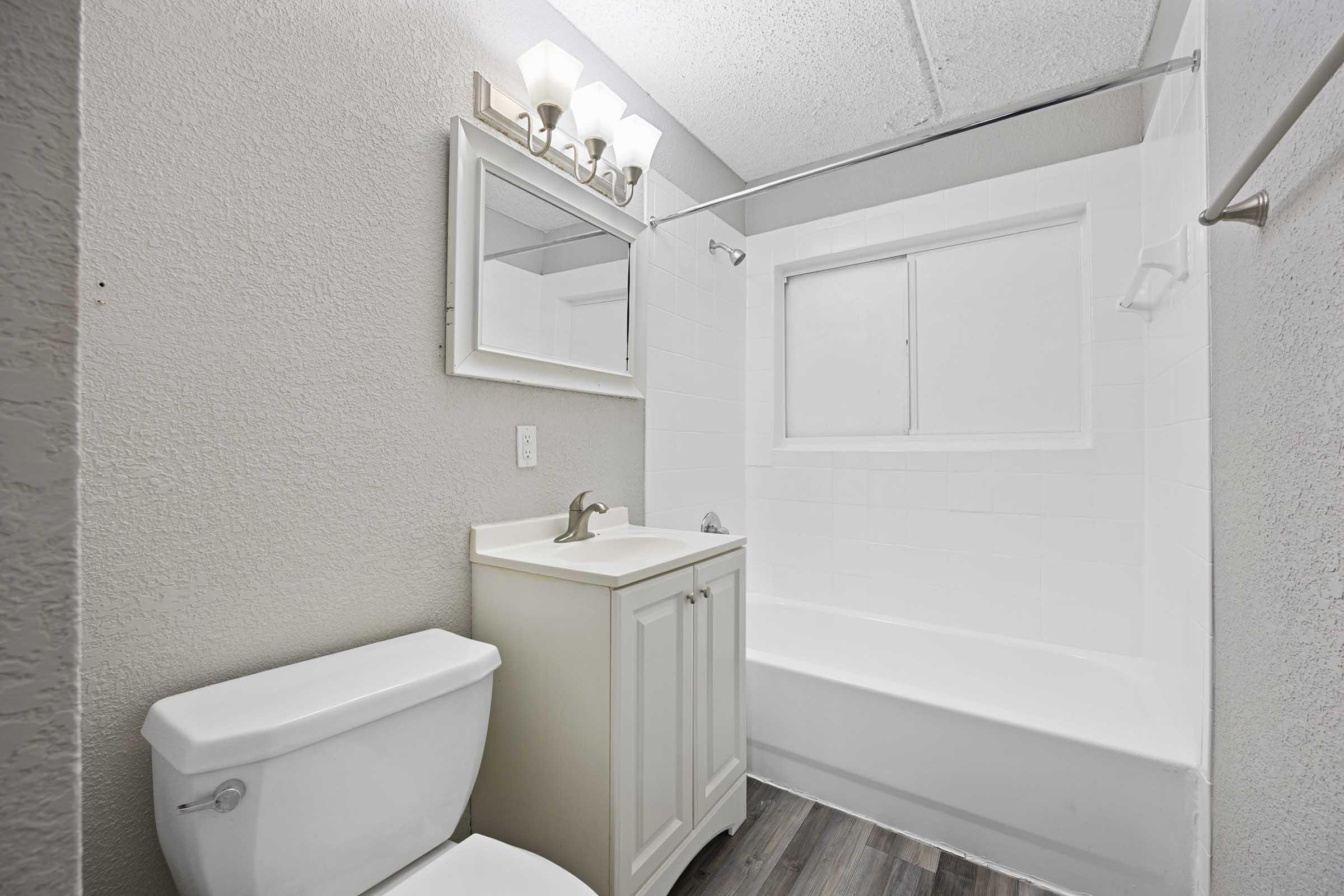 A clean and modern bathroom featuring a white bathtub with a shower, a mirror above a vanity with a sink, and a toilet. The walls are painted in a light color, and there is soft lighting from fixtures above the mirror. The flooring appears to be a wood-like laminate.