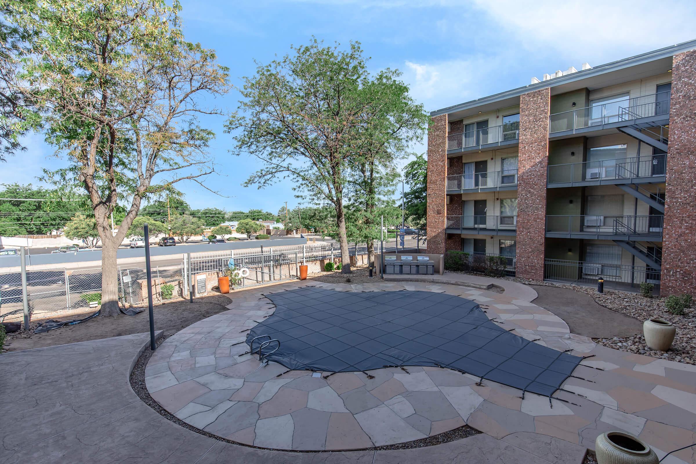 Outdoor courtyard area with a pool cover in the center, surrounded by landscaped stones and potted plants. In the background, a multi-story residential building and a clear blue sky. Trees provide some shade, and there is a fence along the side of the property.