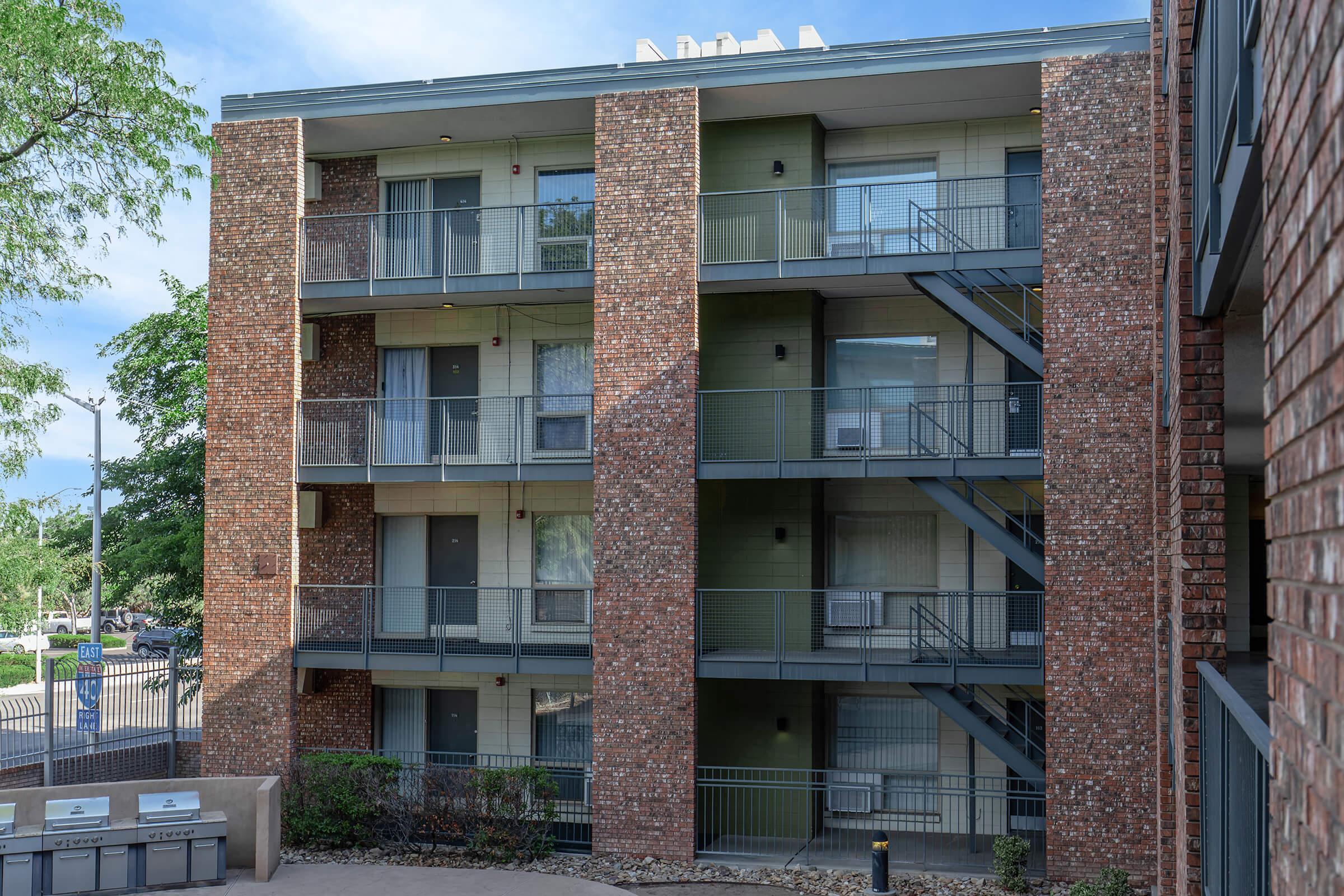 Exterior view of a multi-story brick apartment building featuring multiple balconies with metal railings, outdoor staircases, and a surrounding landscaped area. The scene is set under a clear blue sky, showcasing a well-maintained residential environment.
