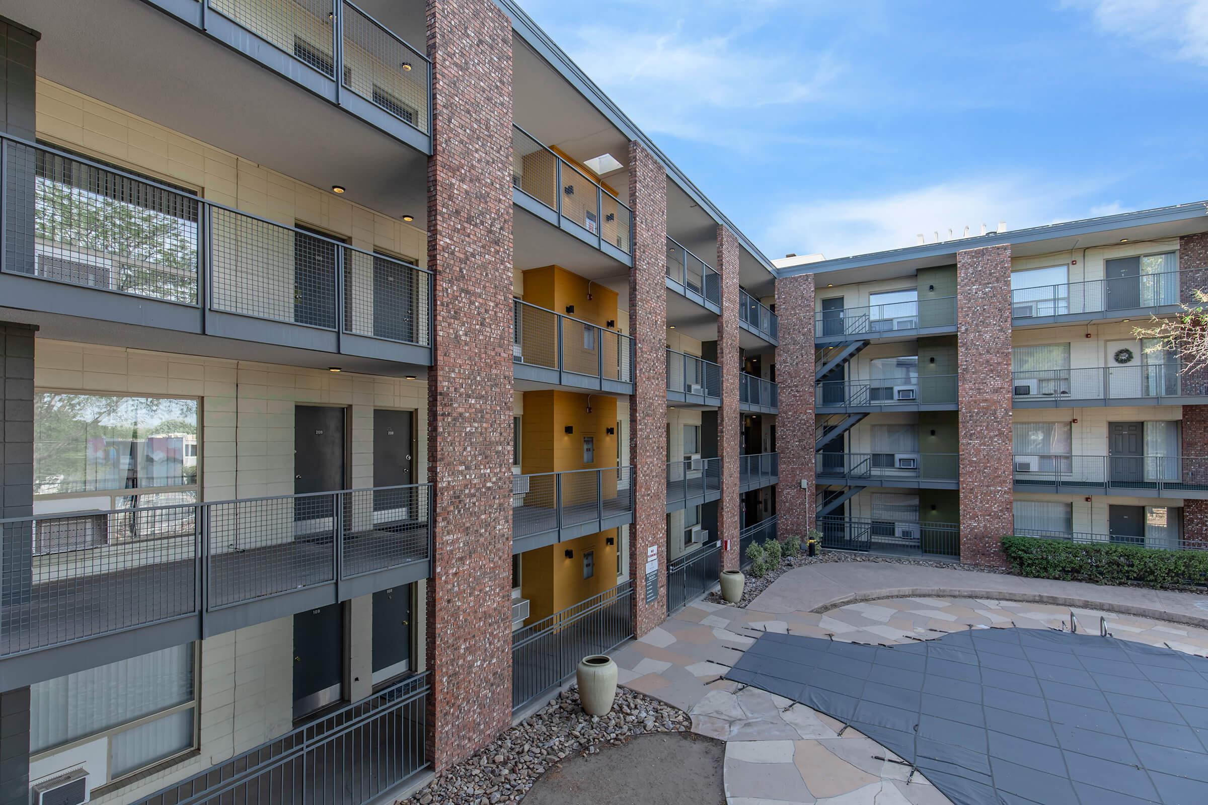 A view of a multi-story apartment complex with brick facades and balconies. The central courtyard features a tiled surface and greenery. The sky is clear, creating a bright atmosphere. Balconies are visible on each level, with doors leading into the units.