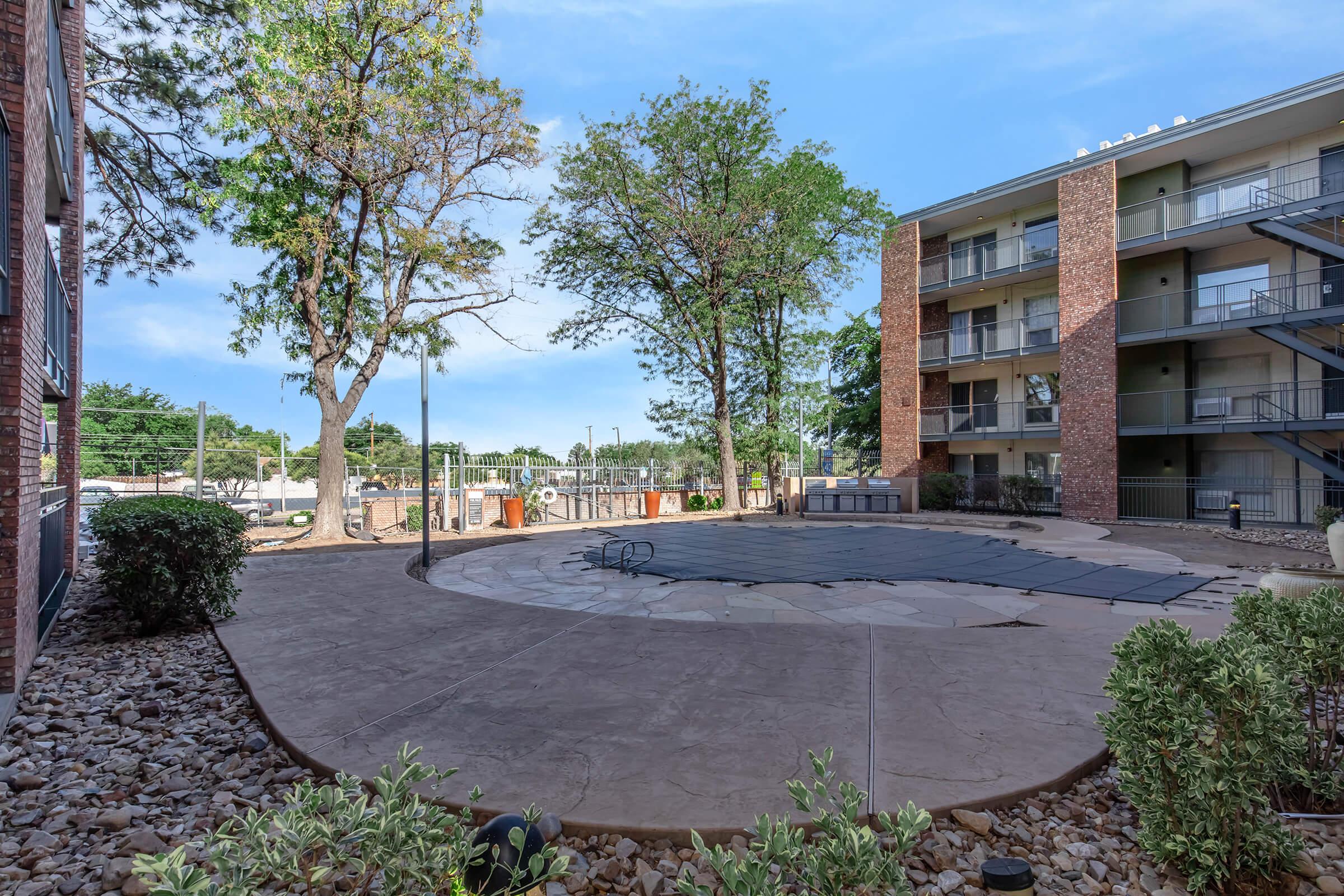 A landscaped outdoor area featuring a pool with a safety cover, surrounded by concrete pathways and trees. Nearby, there are apartment buildings and recreational facilities visible in the background. The scene is well-lit, suggesting a pleasant day.
