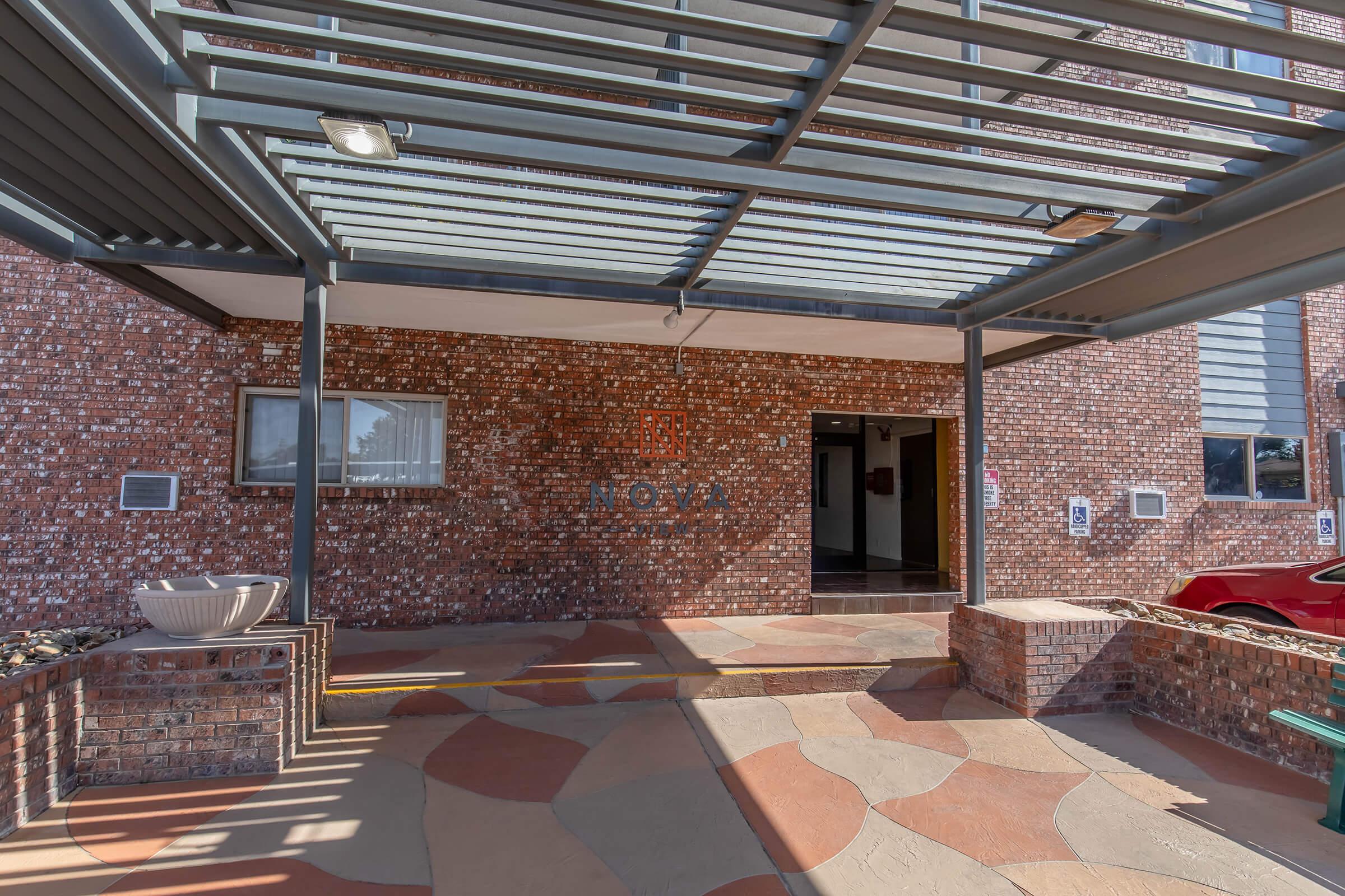 Entrance to a building featuring a decorative brick facade and a covered walkway. There is a small water feature on the left and a red vehicle parked nearby. The area is well-lit with overhead lighting and has a patterned stone pathway leading to the door.