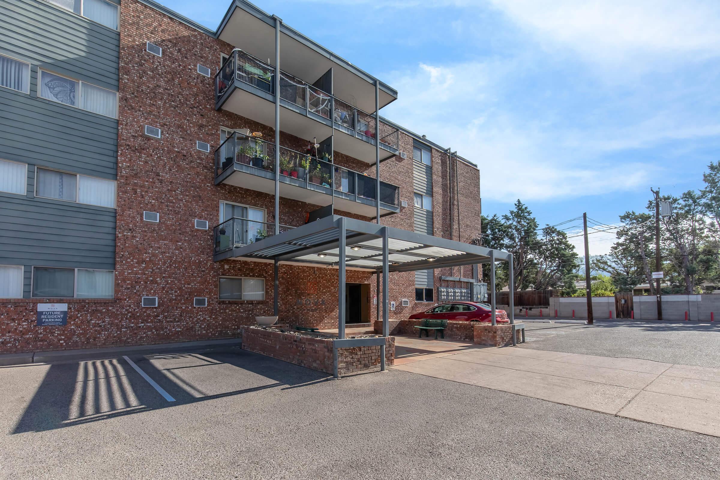 A modern brick apartment building with multiple balconies and a covered entrance area. The parking lot is visible in front, with a few parked cars. The sky is clear and blue, and surrounding trees are present in the background.