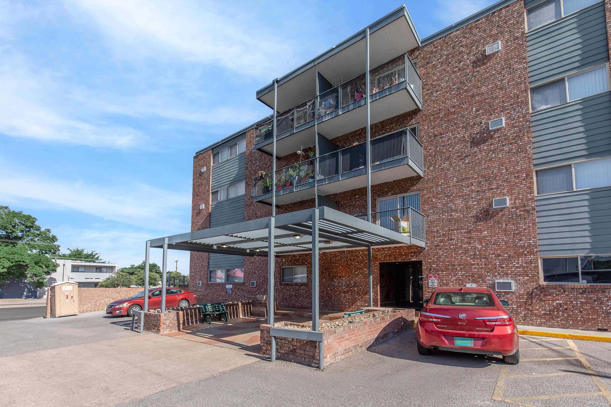 A modern, multi-story brick apartment building featuring several balconies with plants. In the foreground, there are parked cars and a covered entrance area. The sky is clear with scattered clouds, indicating a sunny day. The overall scene conveys a welcoming residential environment.