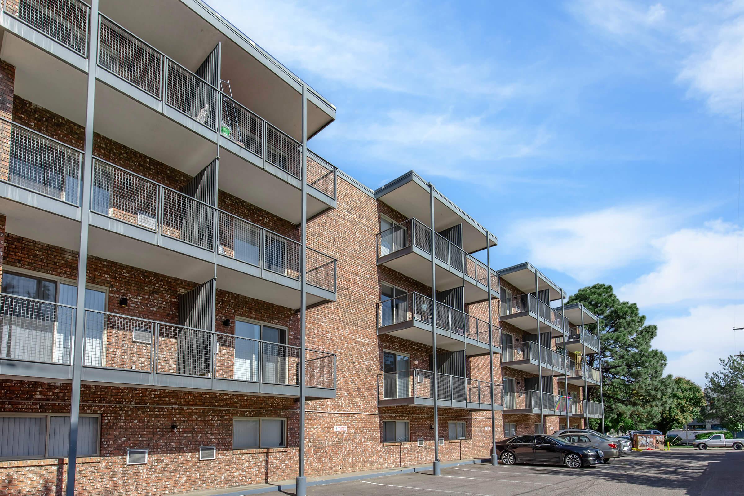 A multi-story brick apartment building featuring several balconies. The building has multiple windows and a modern design. In the foreground, a black car is parked in a lot, with trees and a blue sky in the background.