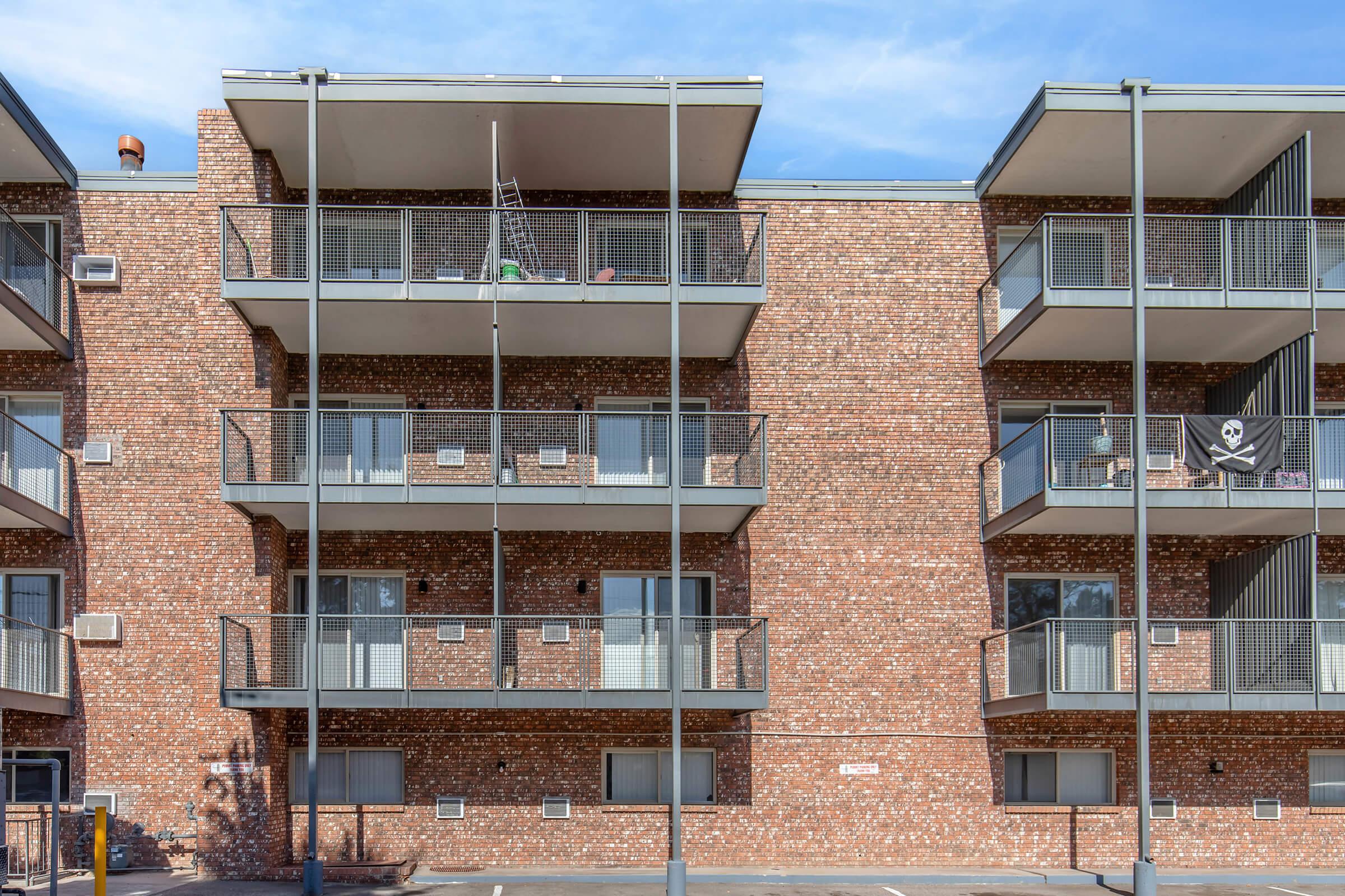 Three-story brick apartment building with multiple balconies and a blue sky overhead. Some balconies have furniture and one displays a pirate flag. The building features a simple architectural design, with windows and metal railings. A parking area is visible in front.