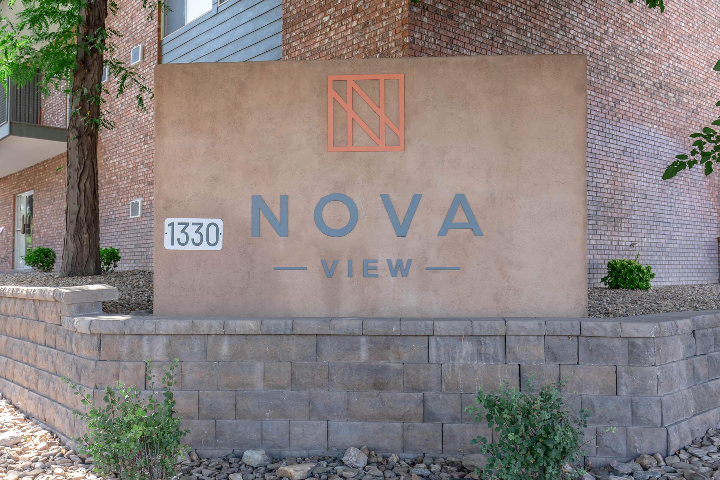 Sign for "NOVA VIEW" located at 1330. The sign features a textured surface with the name "NOVA" in bold blue letters and "VIEW" in smaller text below. An orange logo is positioned above the name. Surrounding the sign are decorative stones and small plants.
