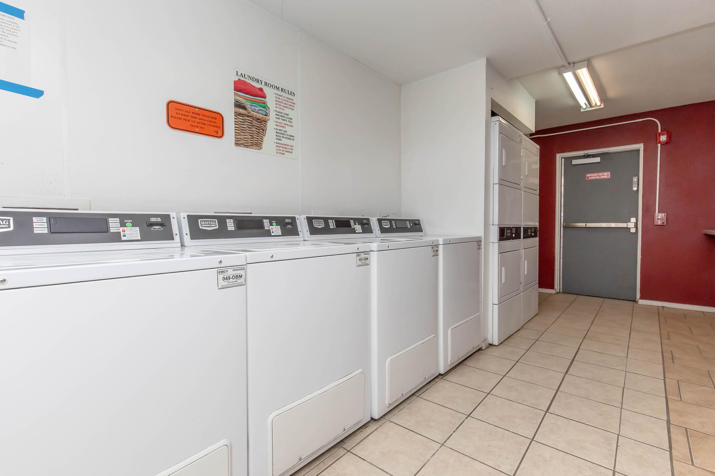 Laundry room with rows of white coin-operated washing machines and dryers against a light-colored wall. A door leads to the outside, and there's a red accent wall with a sign displayed. The floor is tiled, and overhead lighting provides bright illumination.