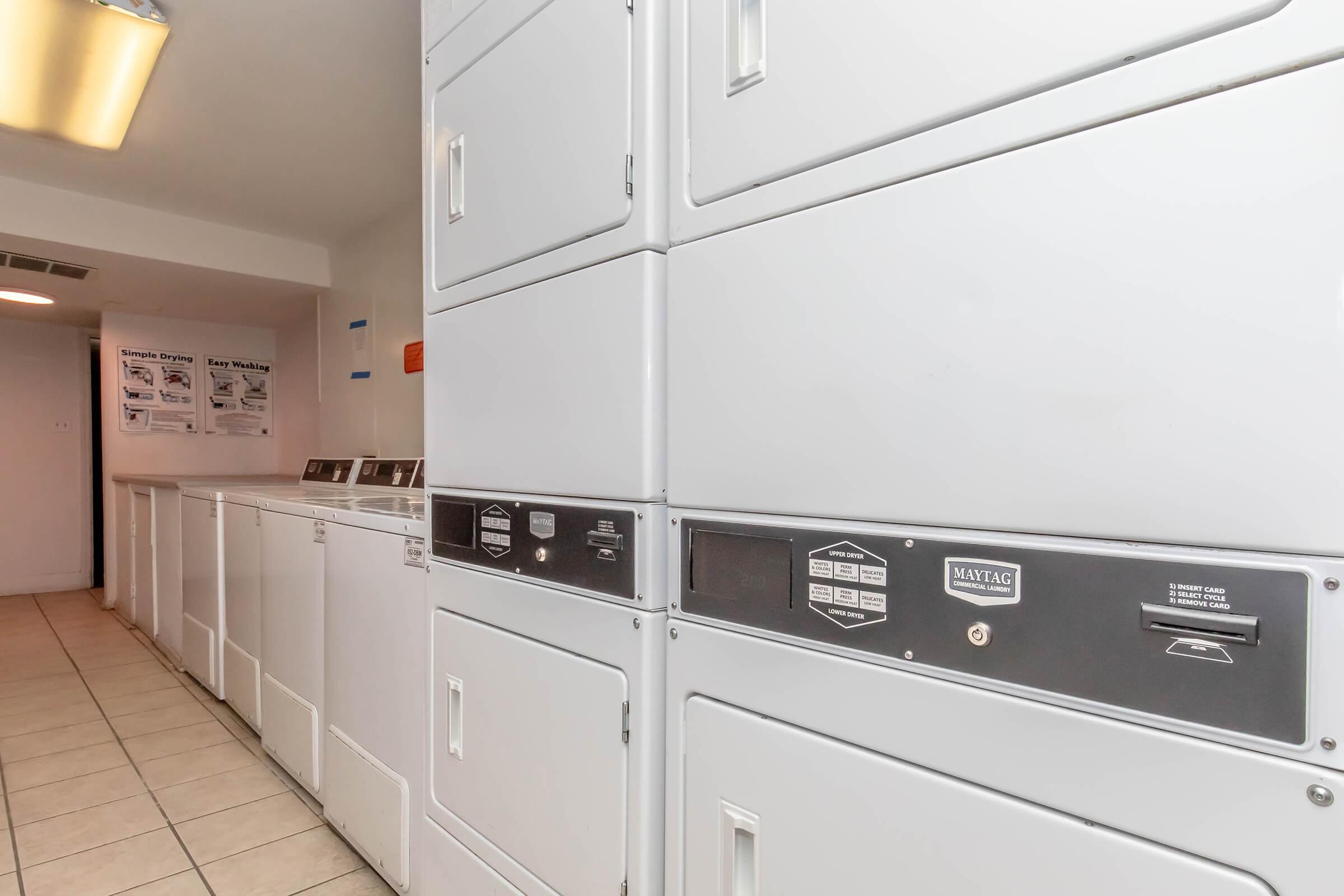 A clean laundry room featuring stacked washers and dryers, along with a row of front-loading machines. The space has tiled flooring and bright overhead lighting, with laundry posters visible on the wall, creating a functional and organized environment for laundry tasks.