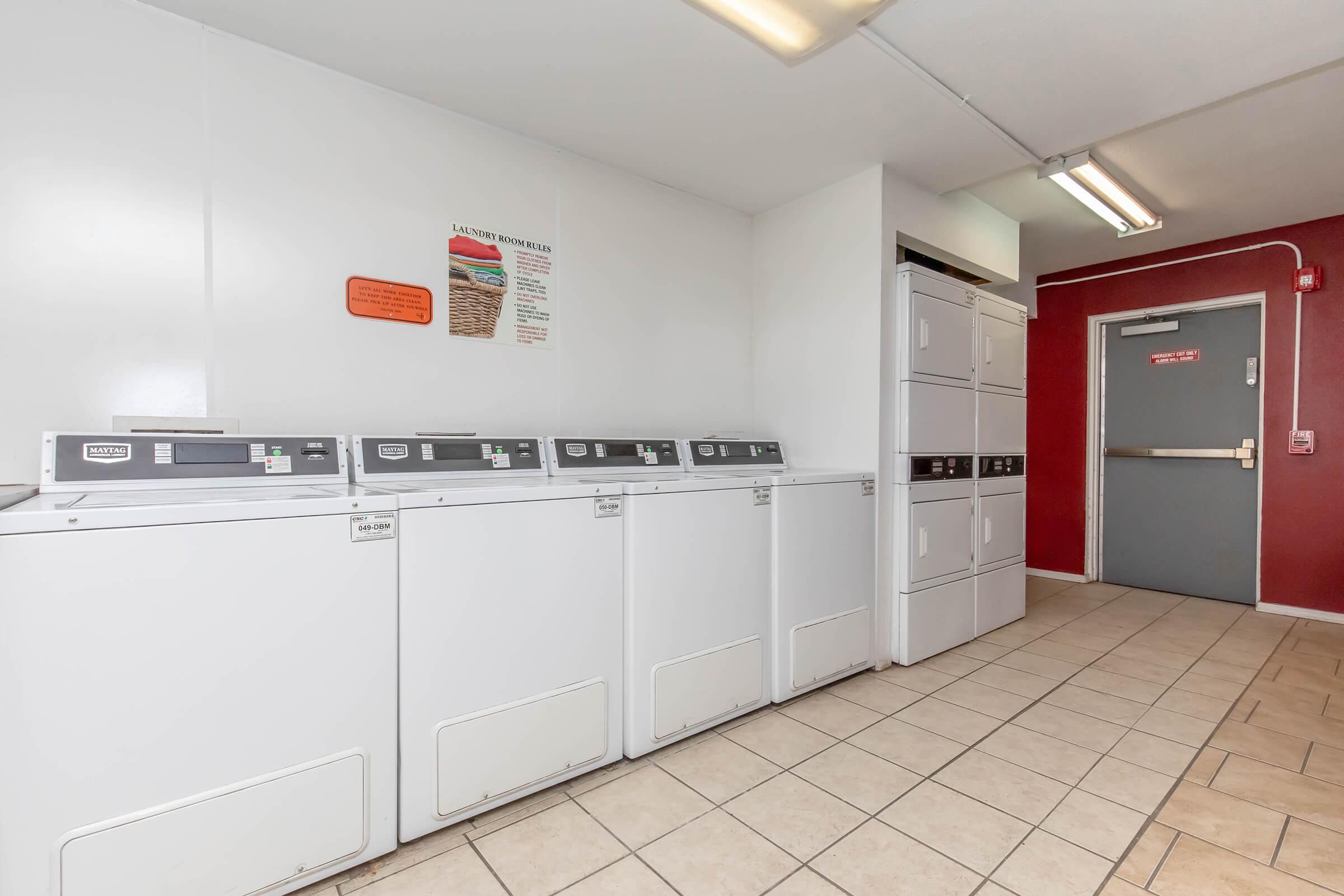 A clean, well-lit laundry room featuring several white washing machines and dryers lined up against a wall. The floor is tiled, and there is a door at the far end of the room. Informational signs are displayed on the wall. The color scheme includes white and red accents.
