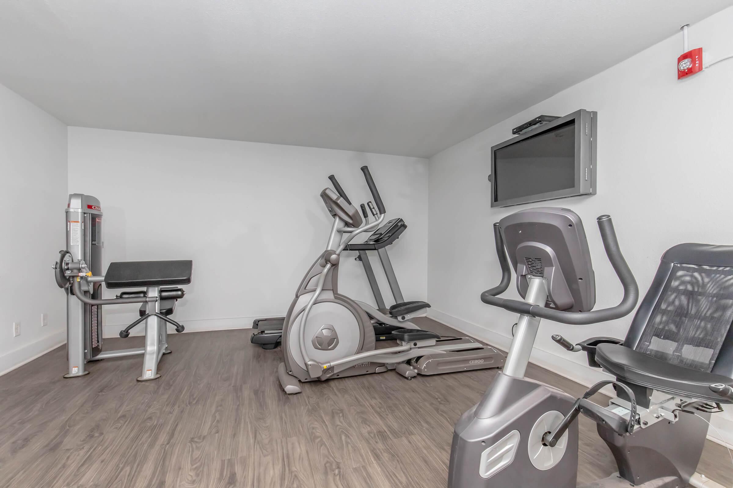 A modern gym space featuring an elliptical machine, a weight bench, and a stationary bike. The room has light-colored walls, wooden flooring, and a wall-mounted television.