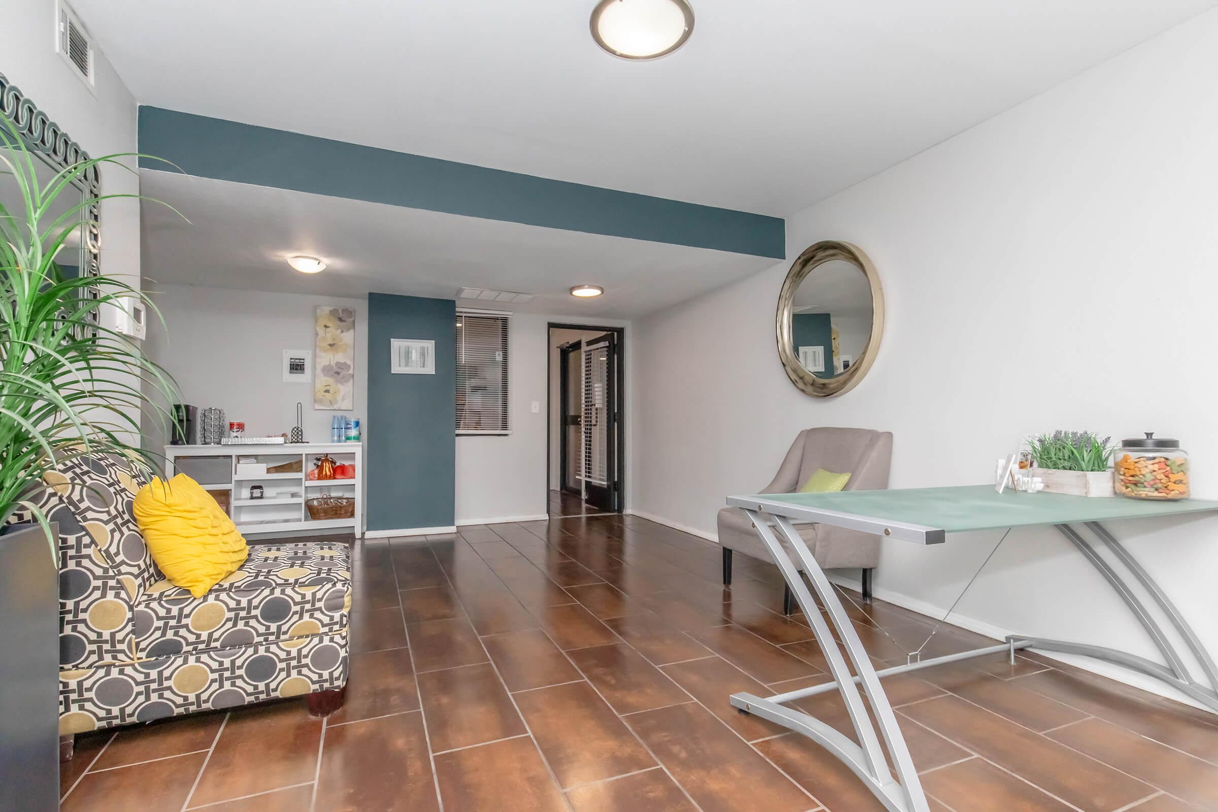 A modern room featuring a patterned armchair with a yellow cushion, a glass-top desk with a small plant and snacks, and a round mirror on the wall. The space has a neutral color palette, elegant lighting, and tiled flooring, creating a bright and inviting atmosphere.