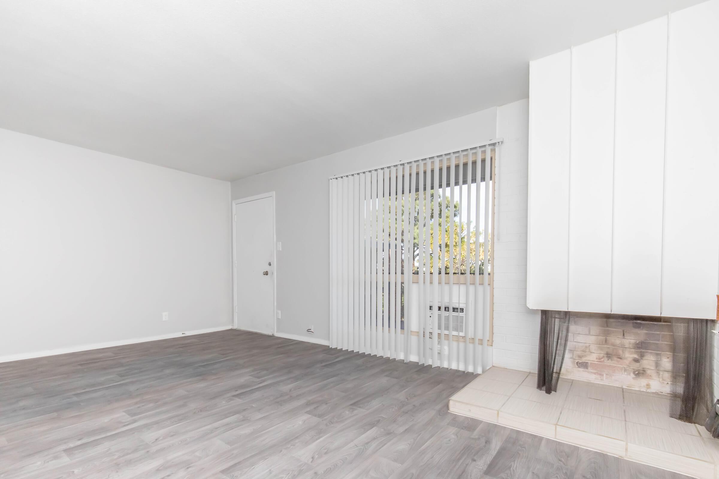 A spacious, empty living room featuring light gray walls, a white door, and large vertical blinds covering a window. A simple fireplace with a tiled base is situated on the right side, and the flooring is a light wood laminate. The space is bright and airy, creating a minimalist atmosphere.